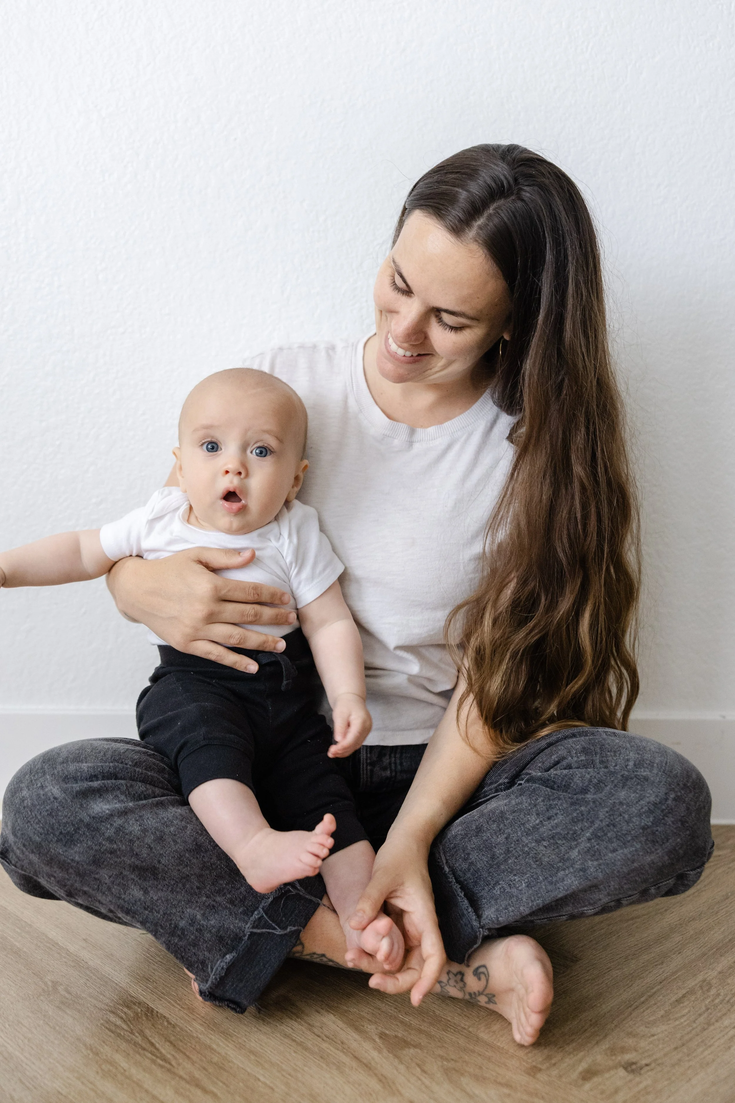 A woman sitting cross-legged on a wooden floor holding a surprised-looking baby in her lap while smiling, against a plain white wall.