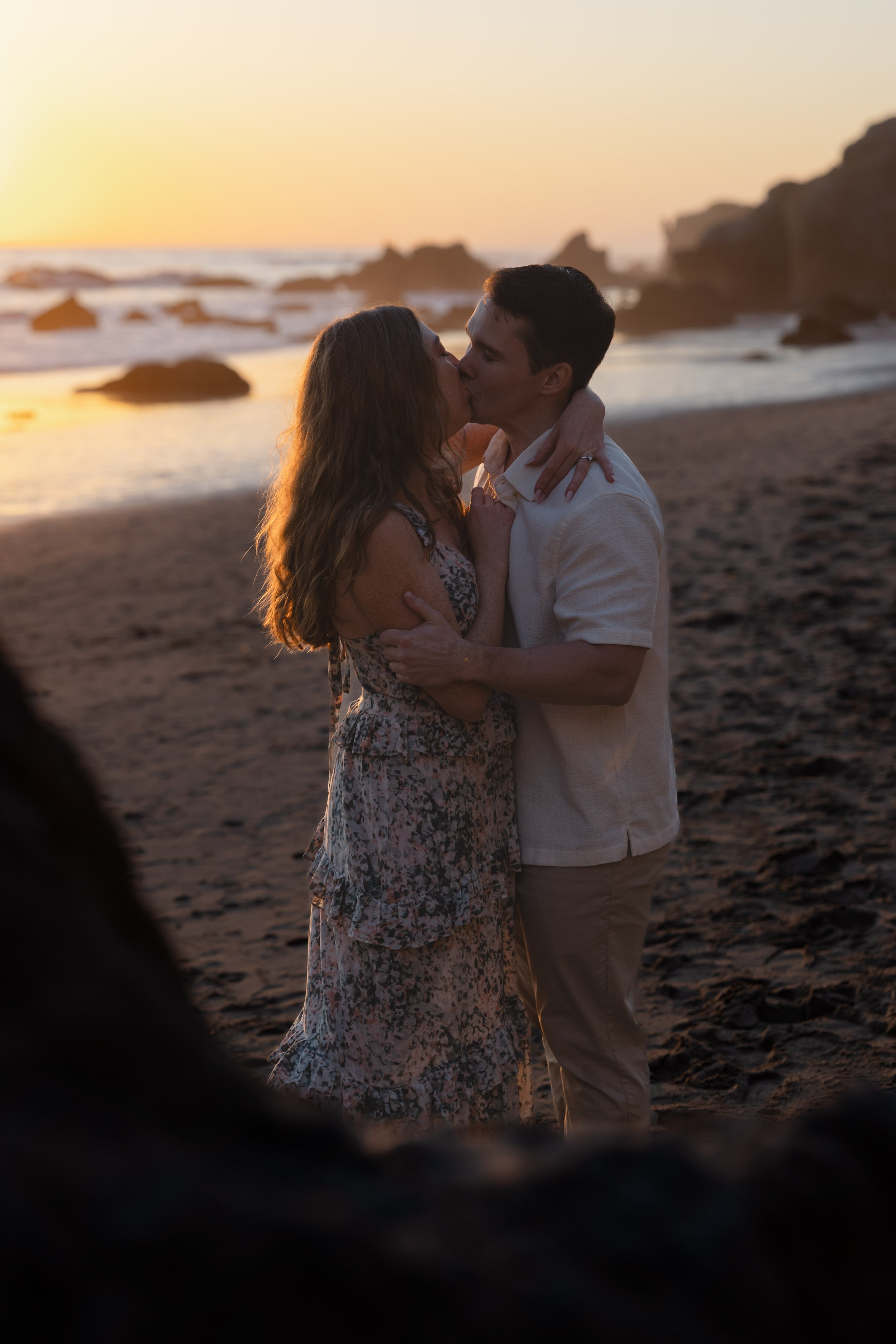 Couple kissing on a beach at sunset, with rocky formations in the background.