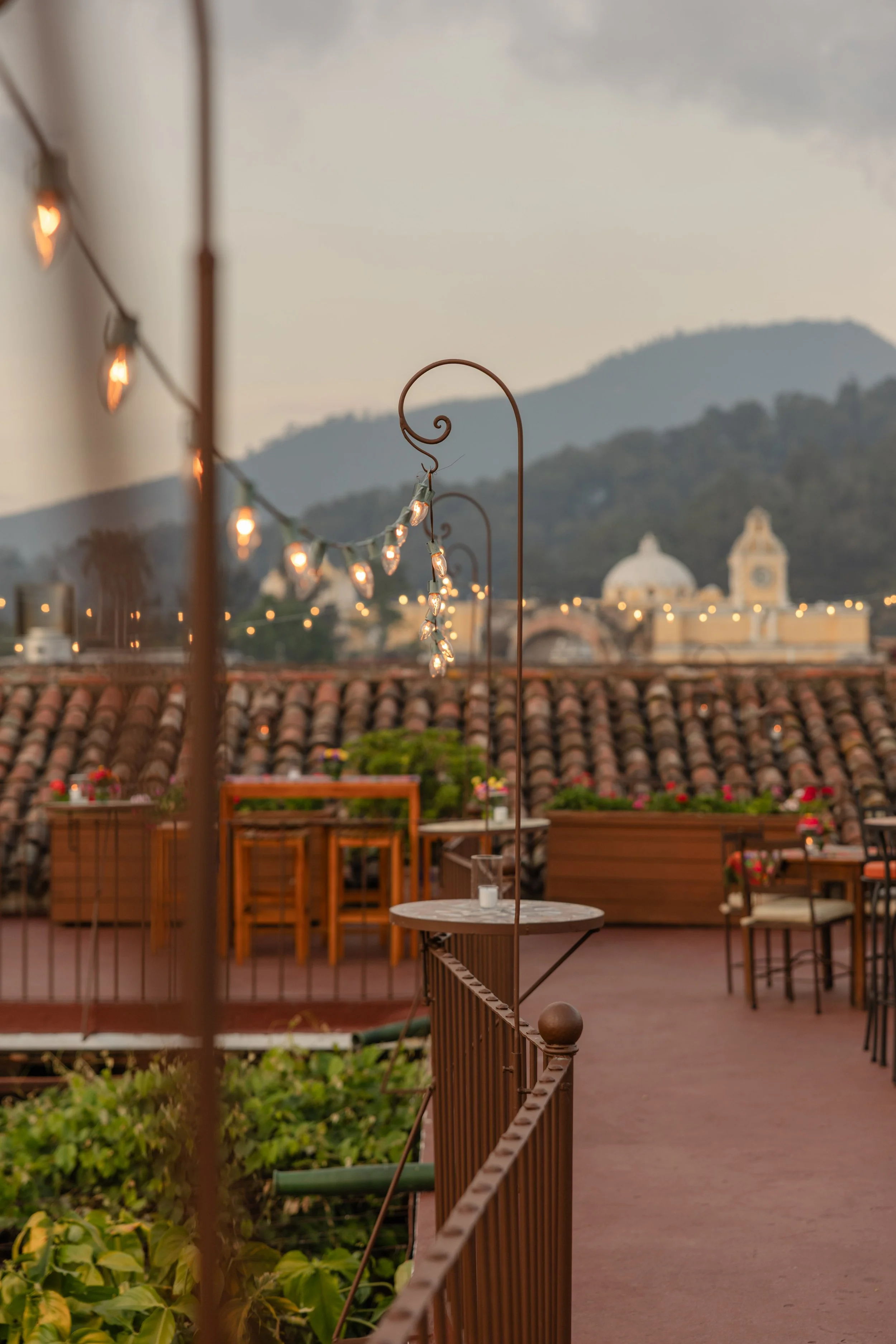 Rooftop terrace with string lights, wooden tables and chairs, potted plants, and a mountain view in the distance at dusk.