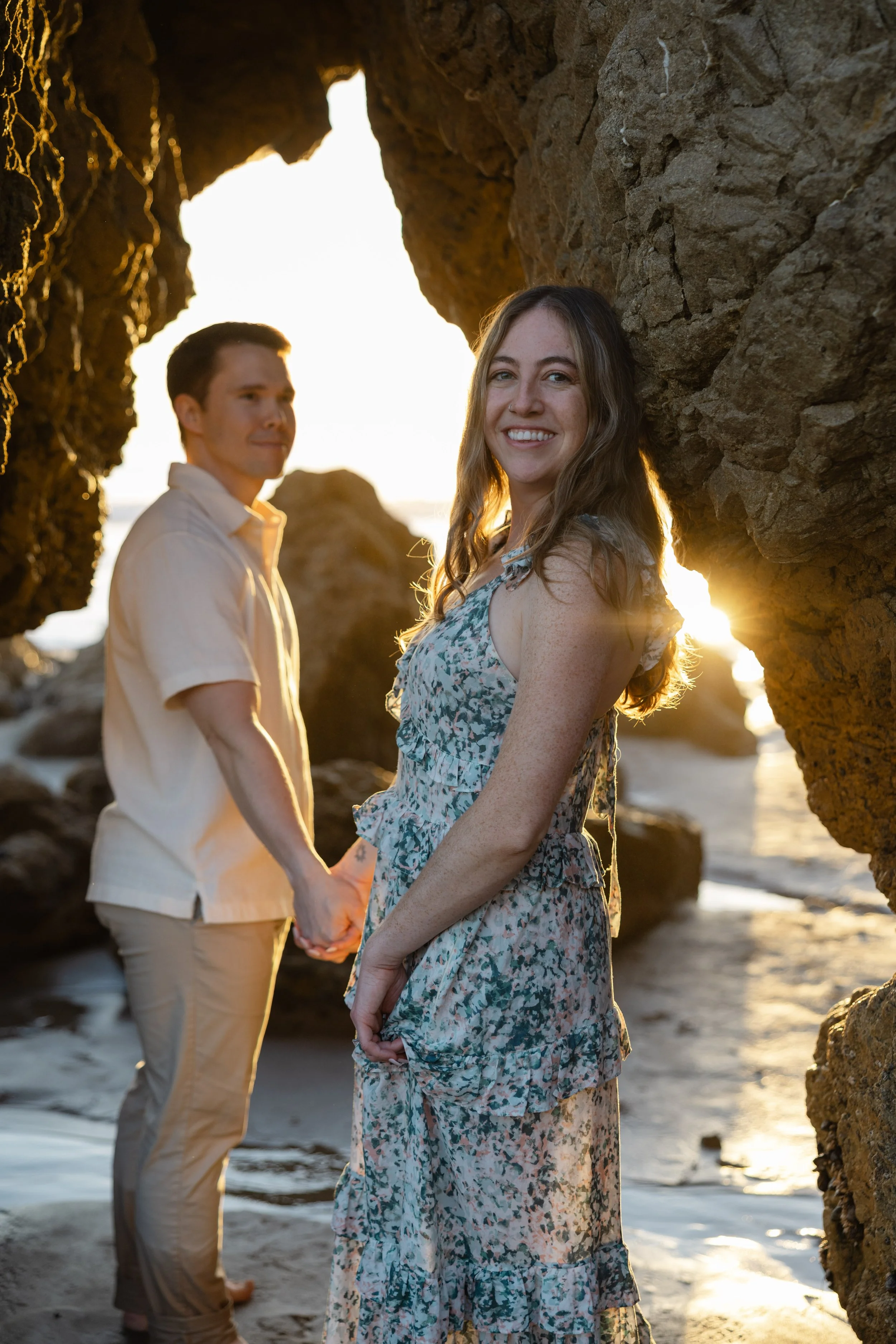 A woman is smiling and holding hands with a man, standing between rock formations during sunset at the beach.