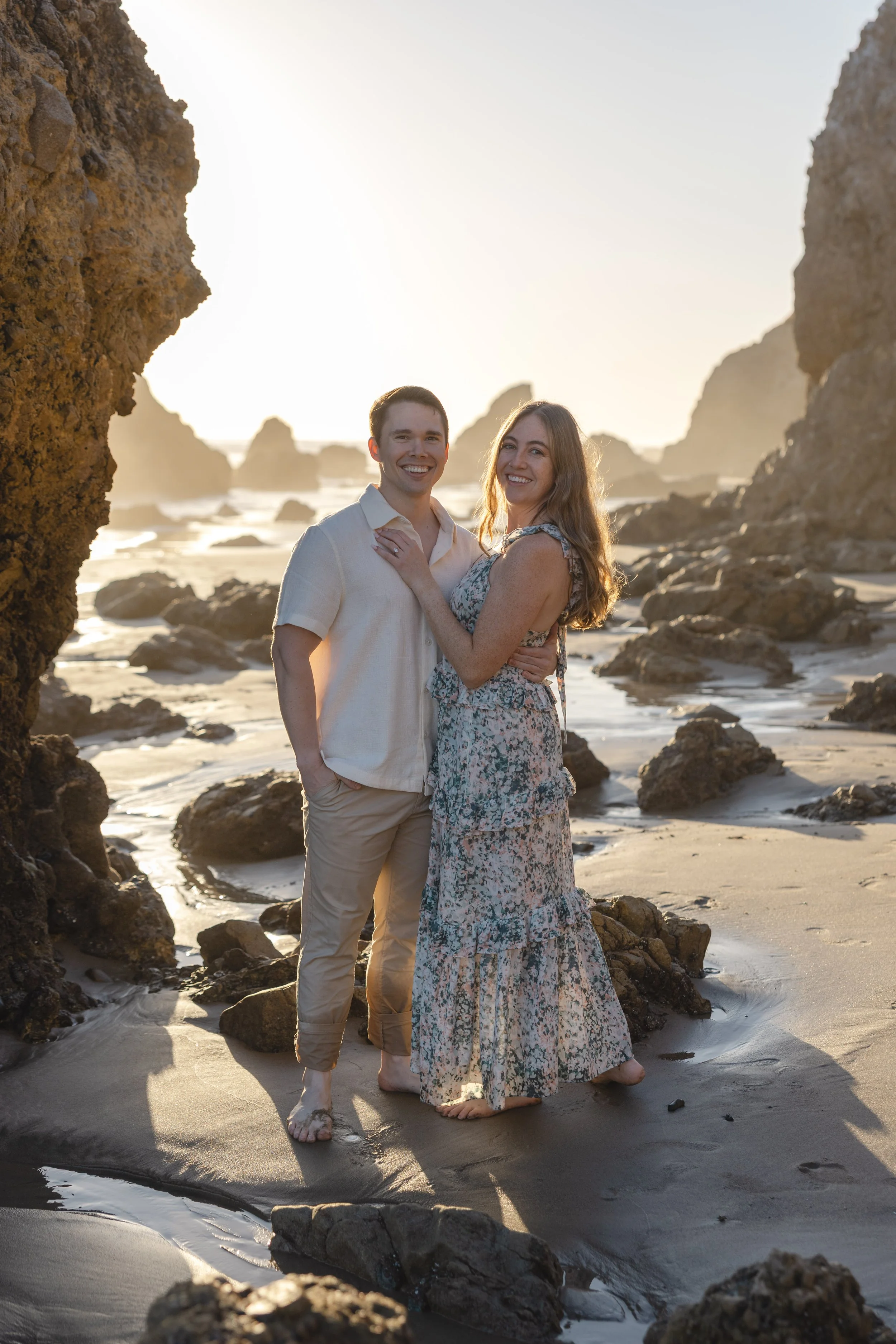 A couple stands on a rocky beach at sunset, smiling and embracing each other.