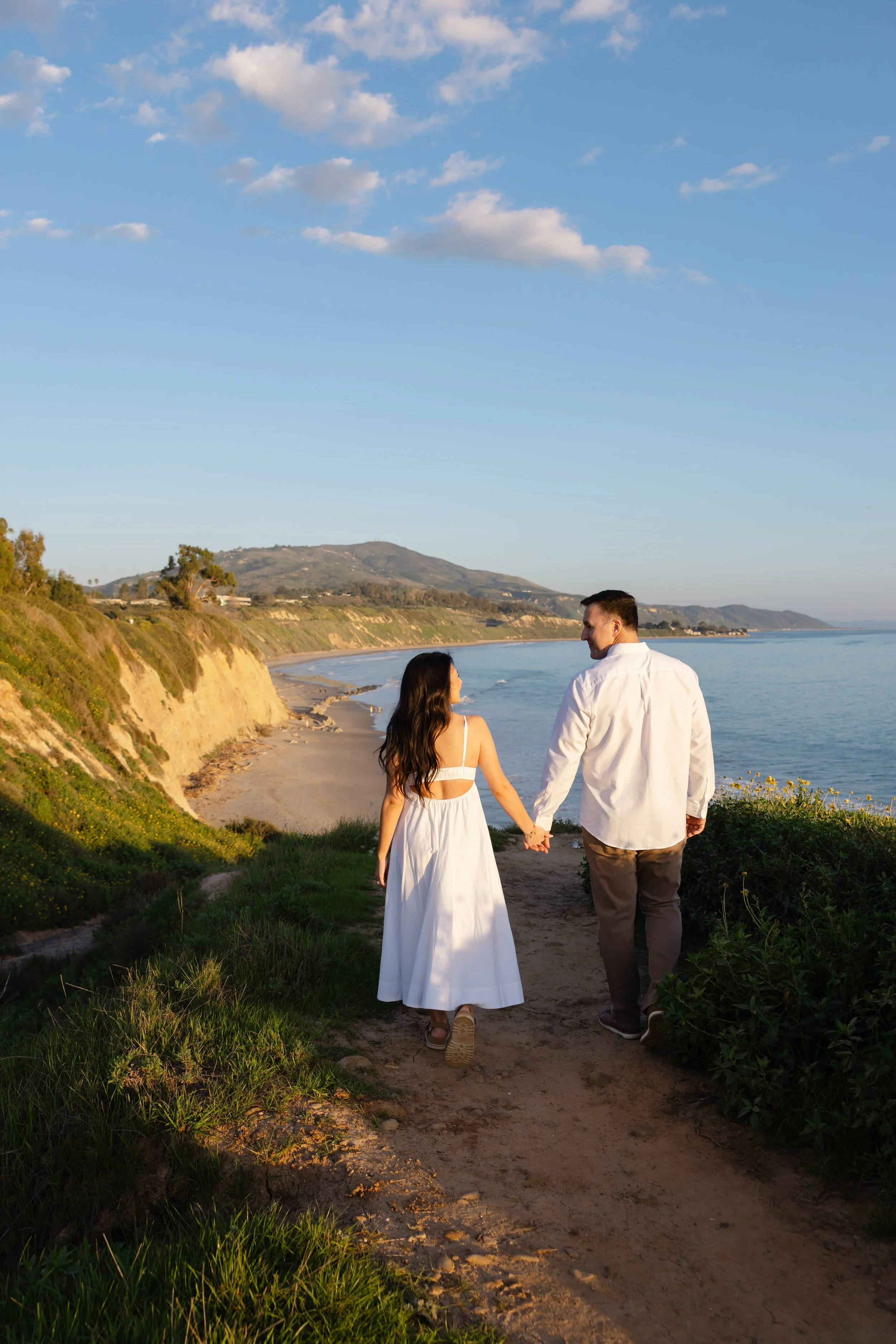 A couple walking holding hands along a scenic coastal pathway with cliffs and the ocean in the background during sunset.