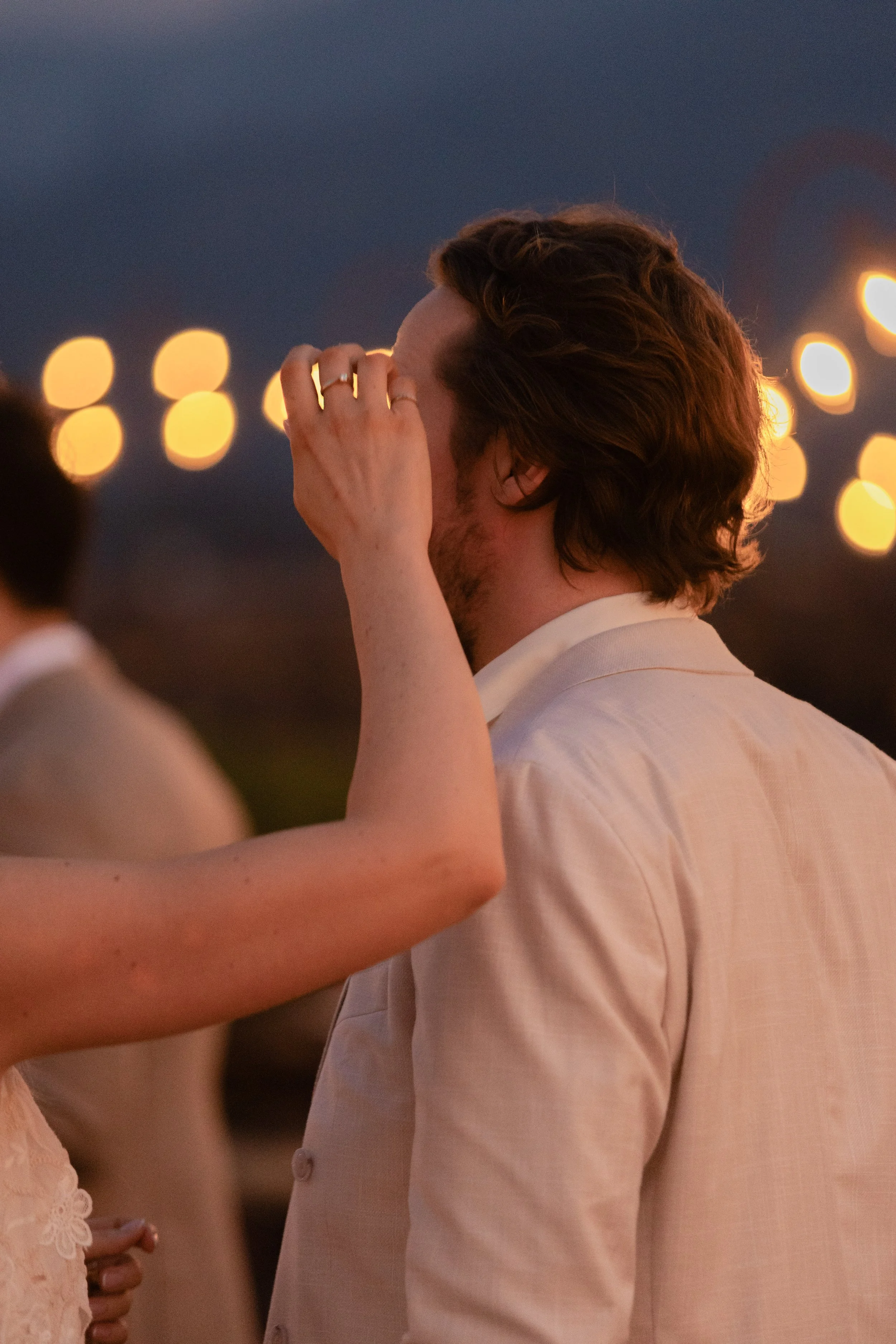 A man with brown hair and a beard wearing a light-colored suit, smiling while covering part of his face with his hand during an outdoor evening event.