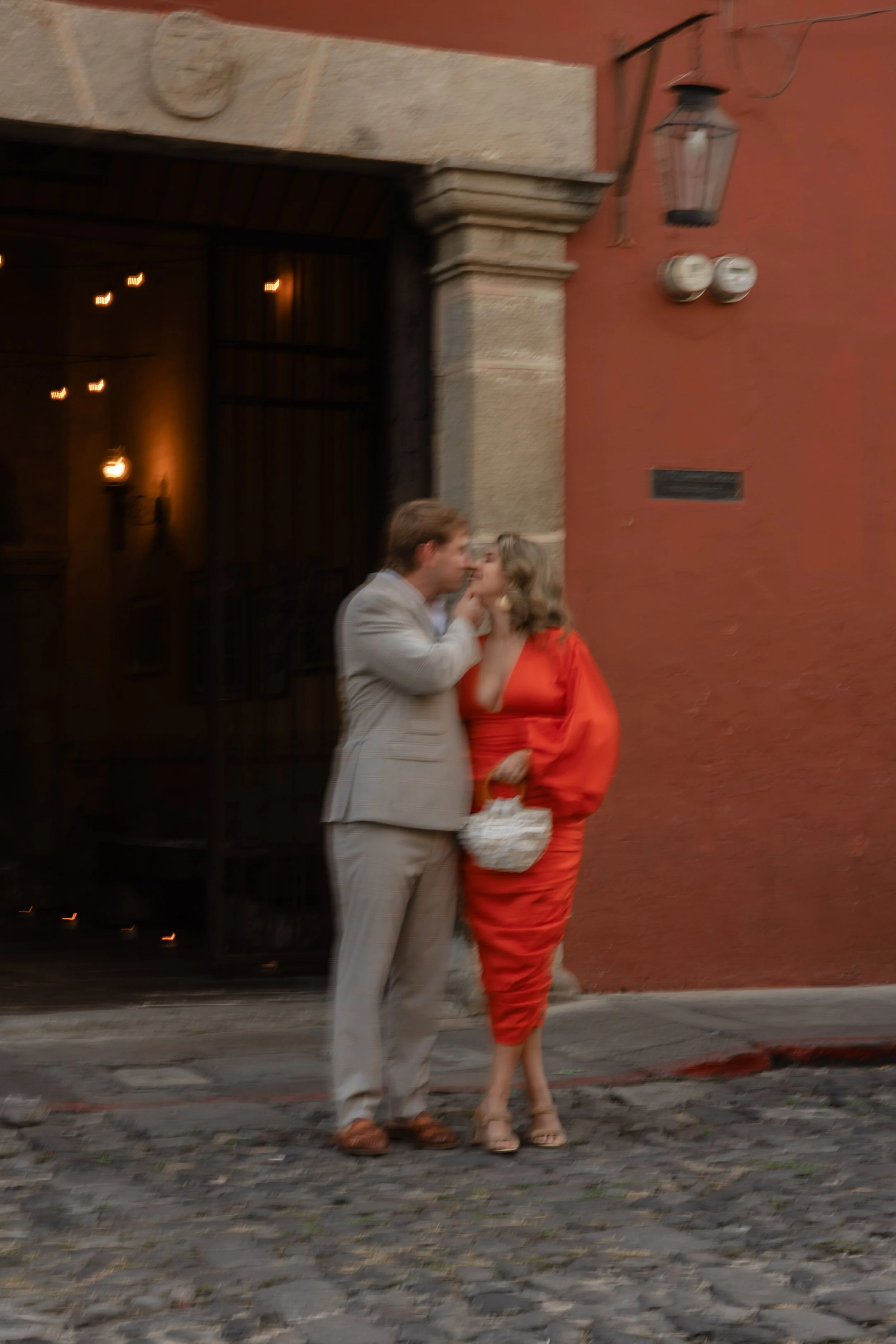 A man and woman share an intimate moment on a cobblestone street outside a building with a reddish wall and vintage lamp. The woman wears a bright red dress, holding a small white purse. The man is in a light-colored suit, leaning toward the woman as