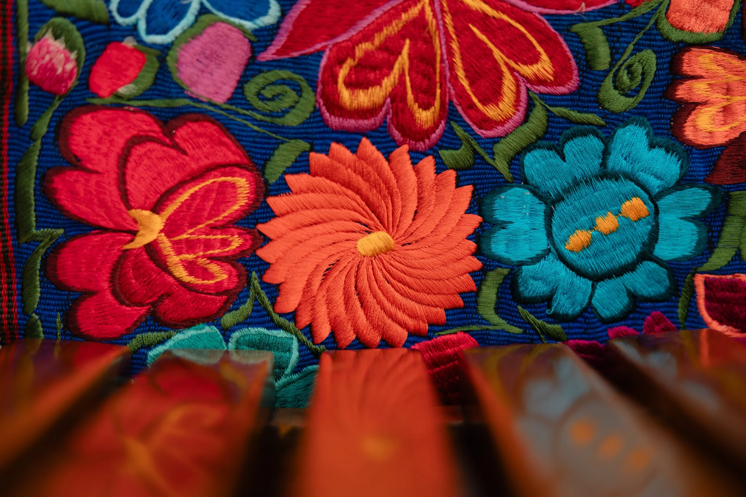 Close-up of colorful embroidered floral pattern on fabric, showing red, orange, turquoise, and pink flowers with green leaves on a blue background.