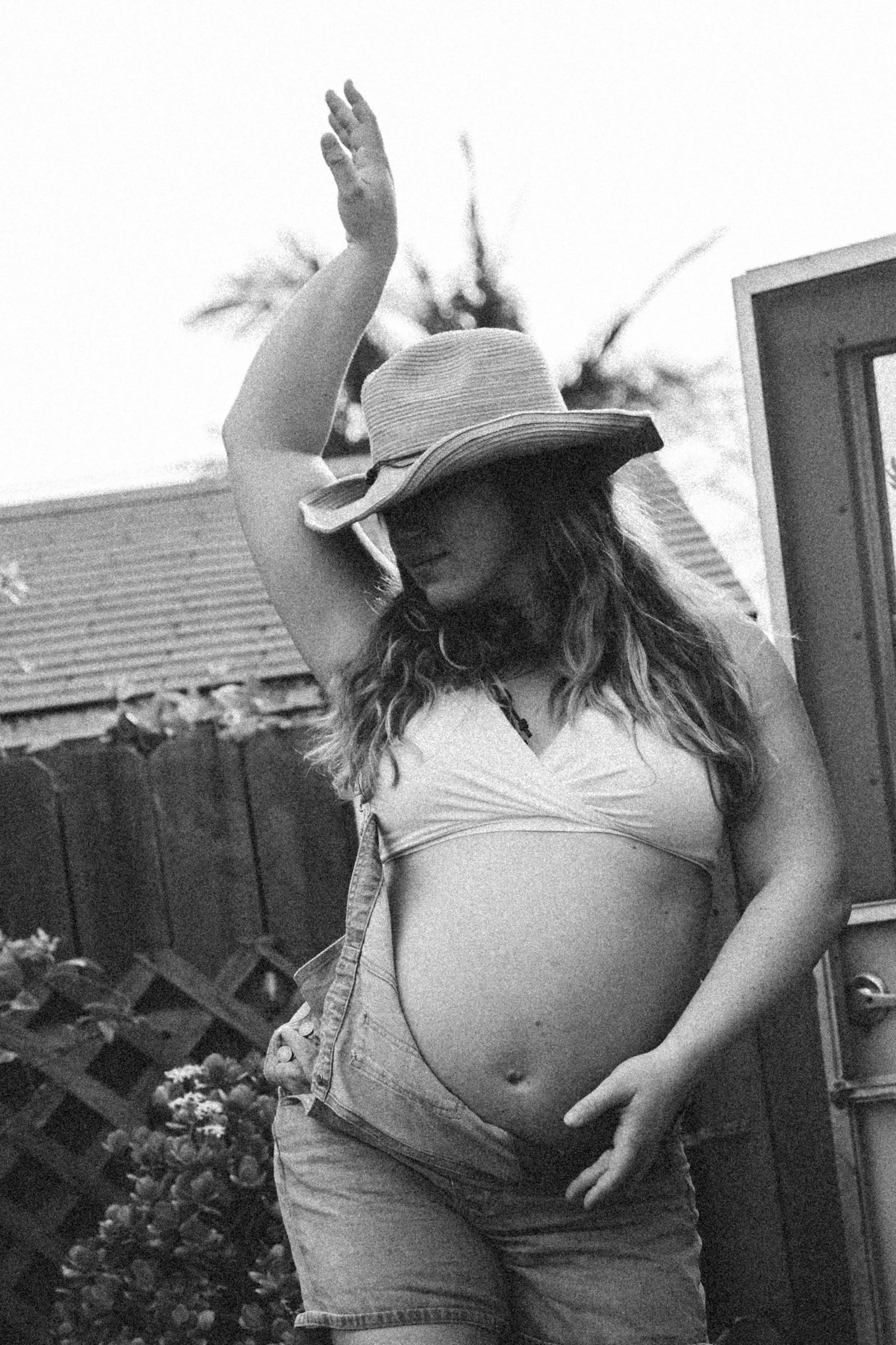 A pregnant woman wearing a hat and a crop top with her hand raised, standing outdoors near a fence, with plants and trees in the background.
