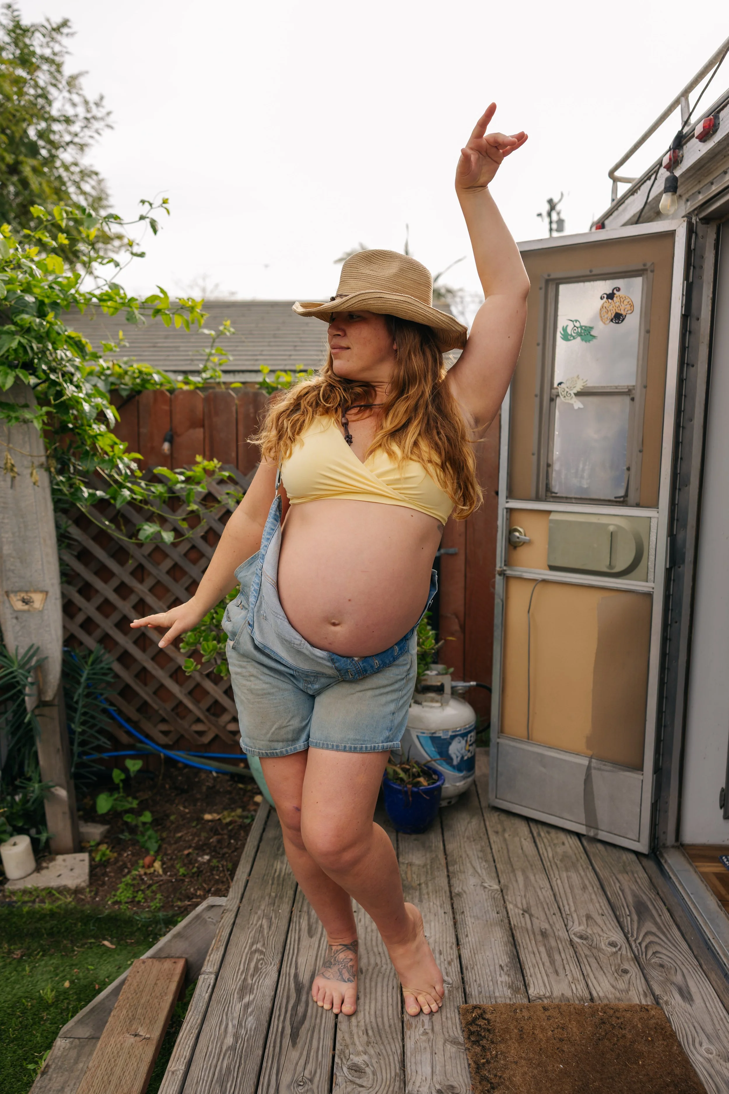 A pregnant woman in a yellow crop top, denim shorts, and a straw hat is dancing on a wooden deck outdoors.