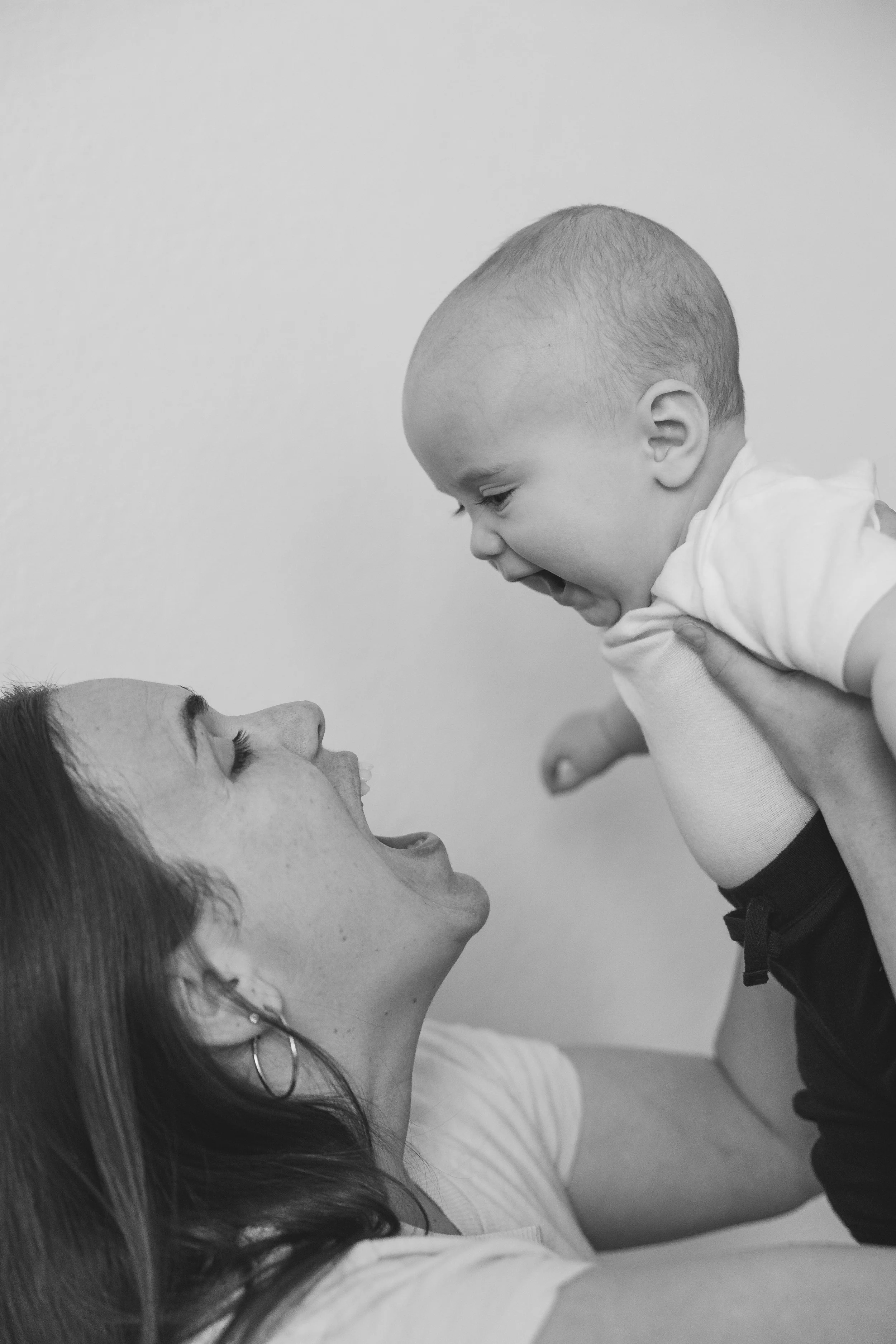 A woman lying on her back holding a baby boy above her. The woman is smiling, and the baby is looking at her with his mouth open. The scene is playful and joyful.