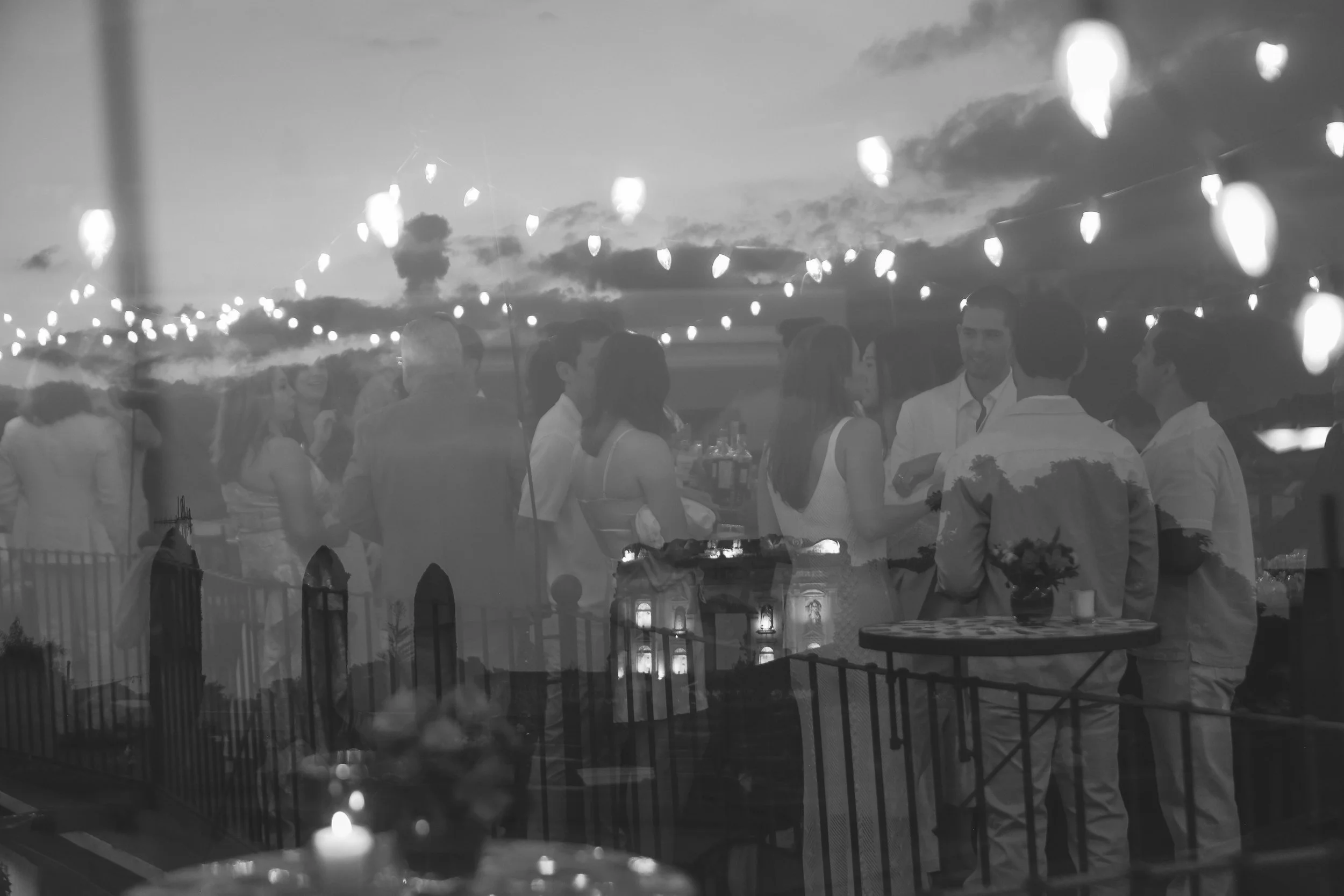 People gathering at an outdoor evening event, viewed through a window with string lights and a candle on a table in the foreground.