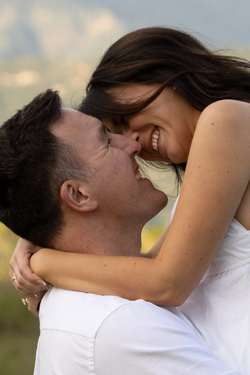 A couple smiling and holding each other closely outdoors, their foreheads touching, in a joyful moment.