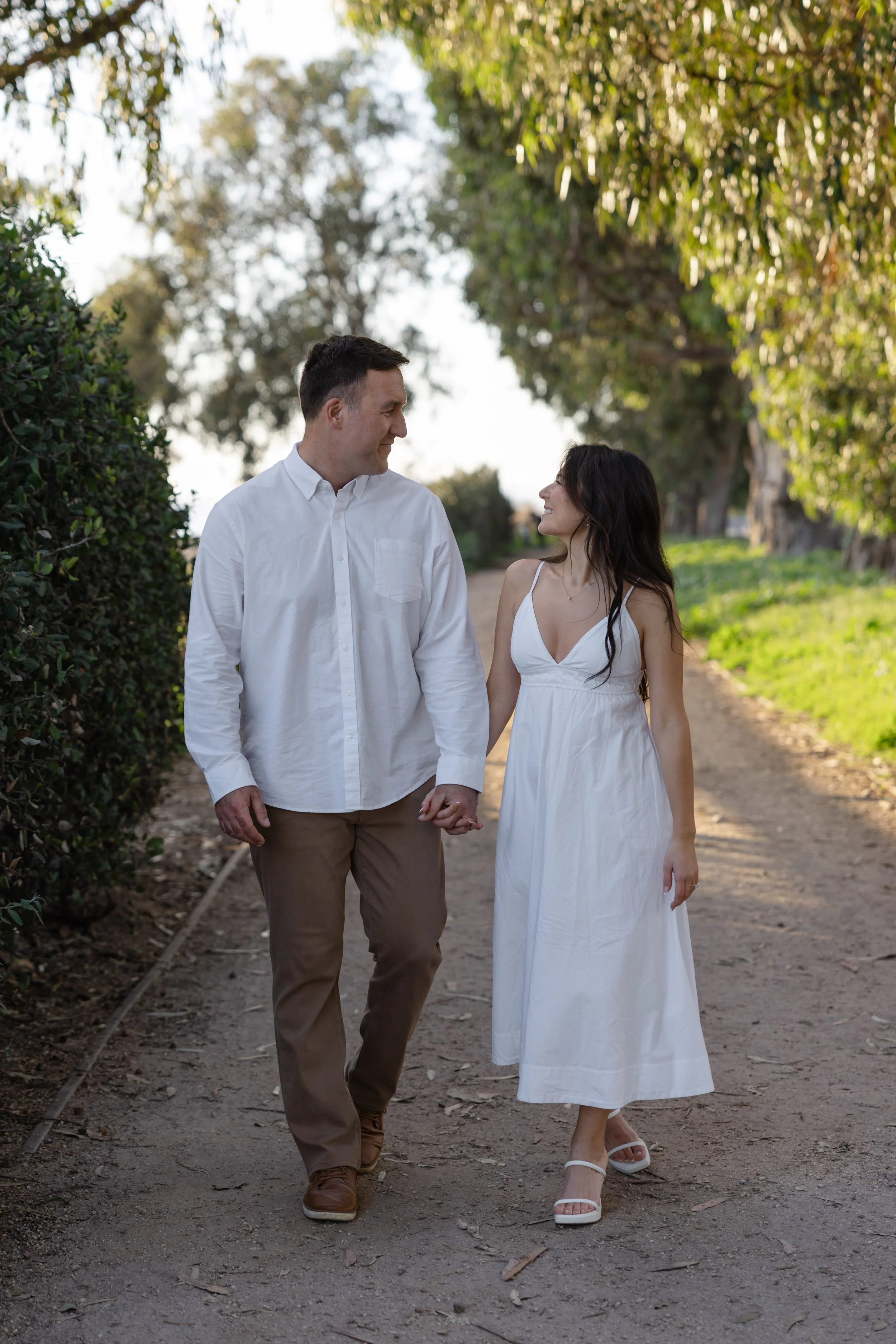 A couple holding hands and walking on a dirt path in a park, with trees and sunlight in the background.