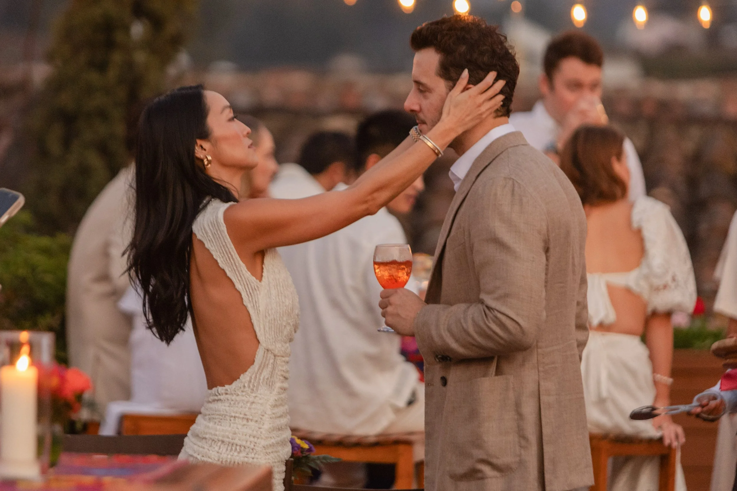 A woman is touching a man's face during a social event outdoors at sunset. The woman has long dark hair and is wearing a sleeveless dress, the man is holding a glass of pink wine and wearing a beige suit. Other people are in the background, some wear