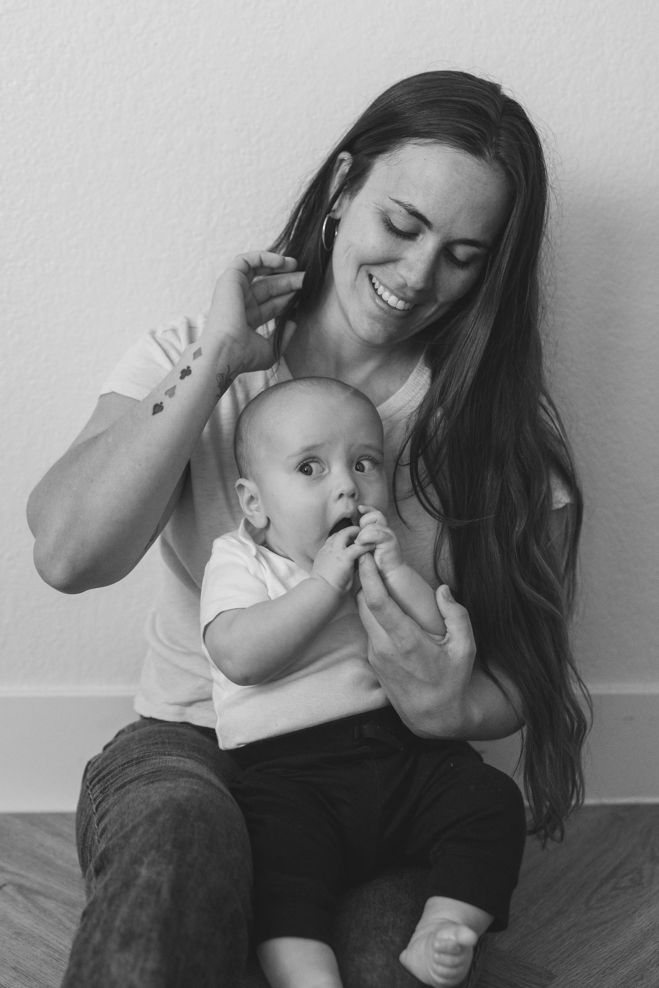 A woman with long hair and tattoos on her arm sitting on the floor, smiling while holding a baby who looks surprised, with a hand in the baby's mouth, against a plain wall.