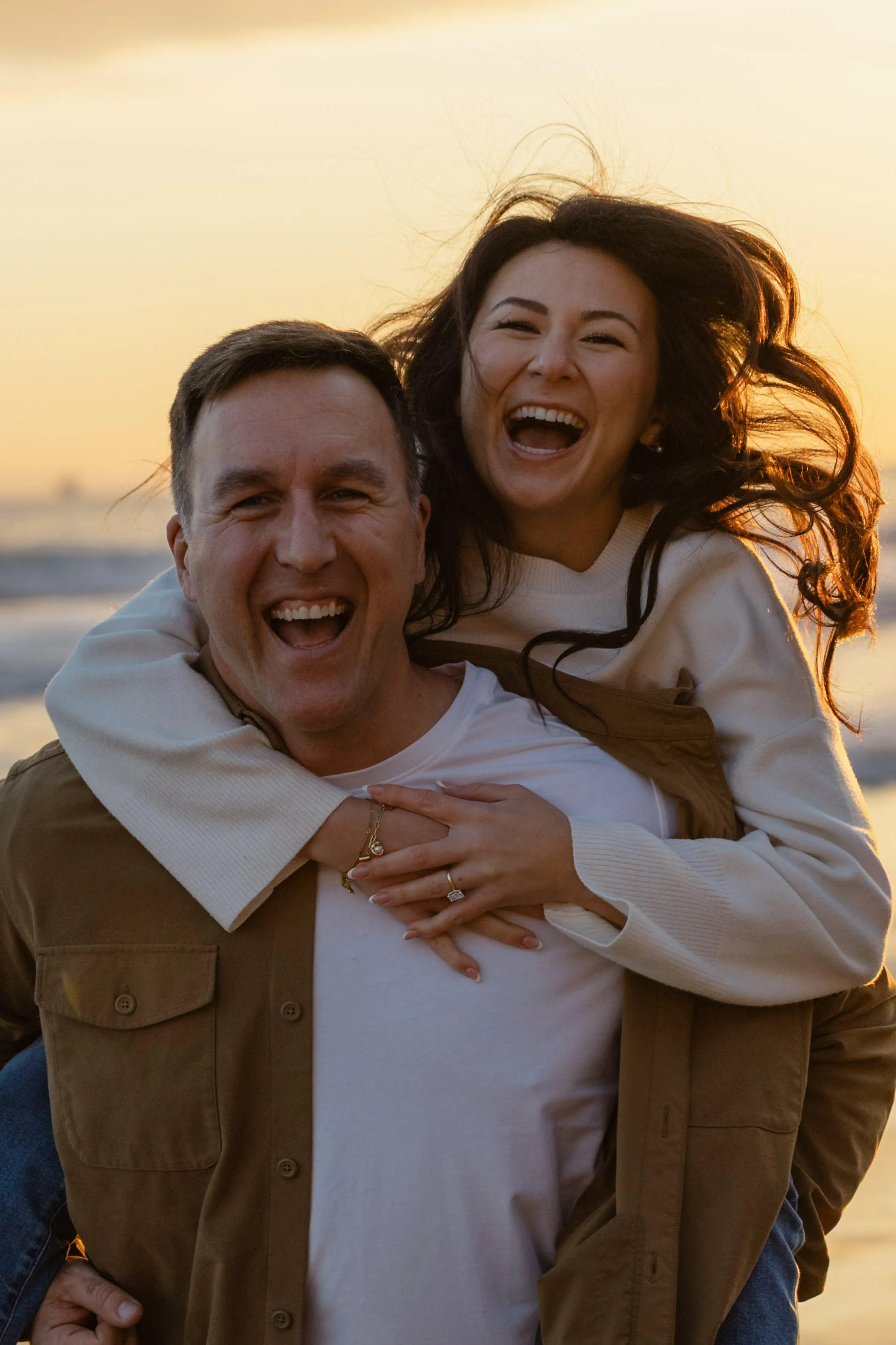A happy couple at the beach during sunset, with the woman piggybacking on the man's back, both laughing.