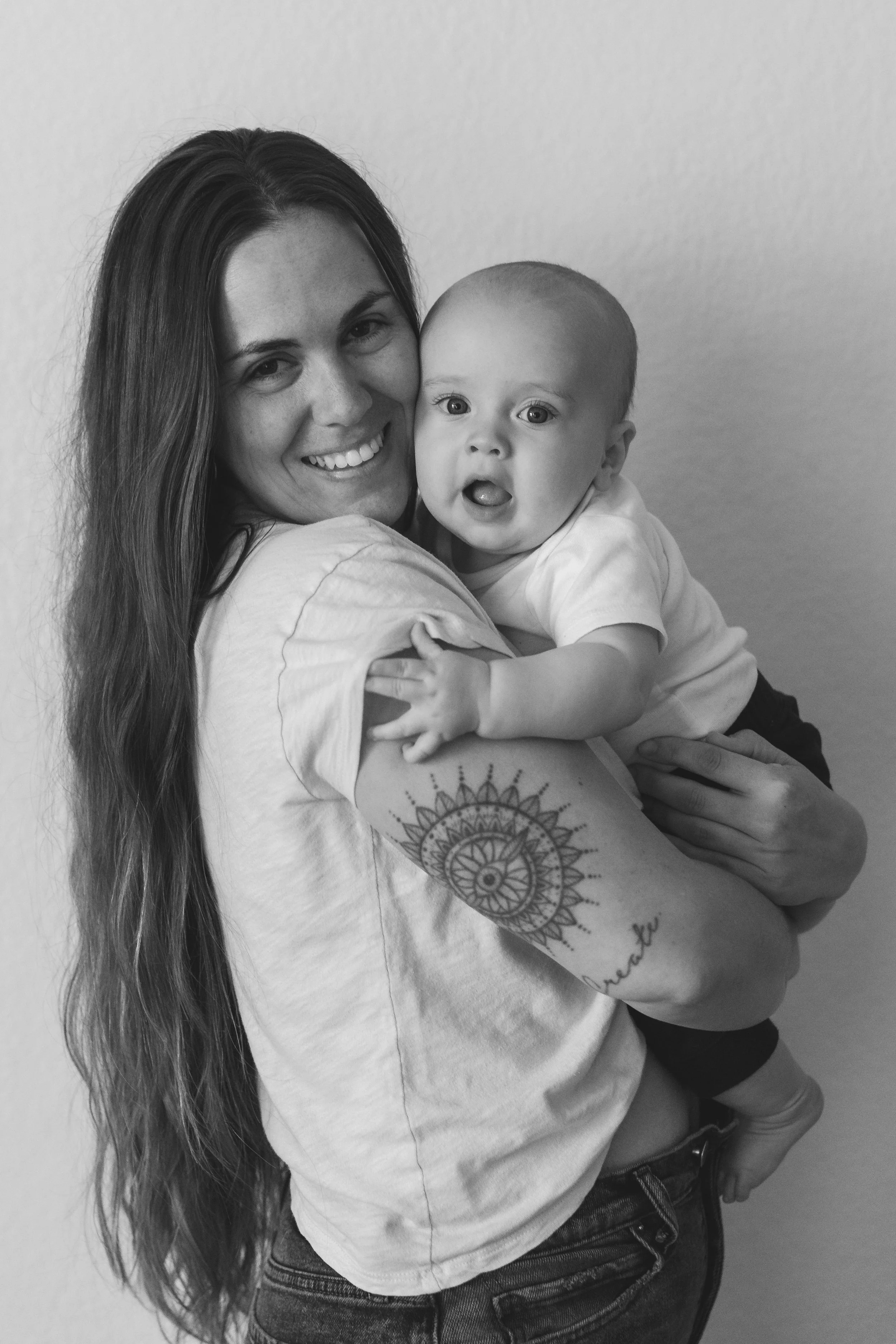A woman with long hair and a tattoo on her arm holding a baby, both smiling, black and white photograph.