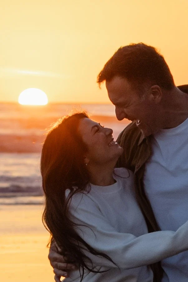A couple embracing on the beach during sunset, smiling at each other.