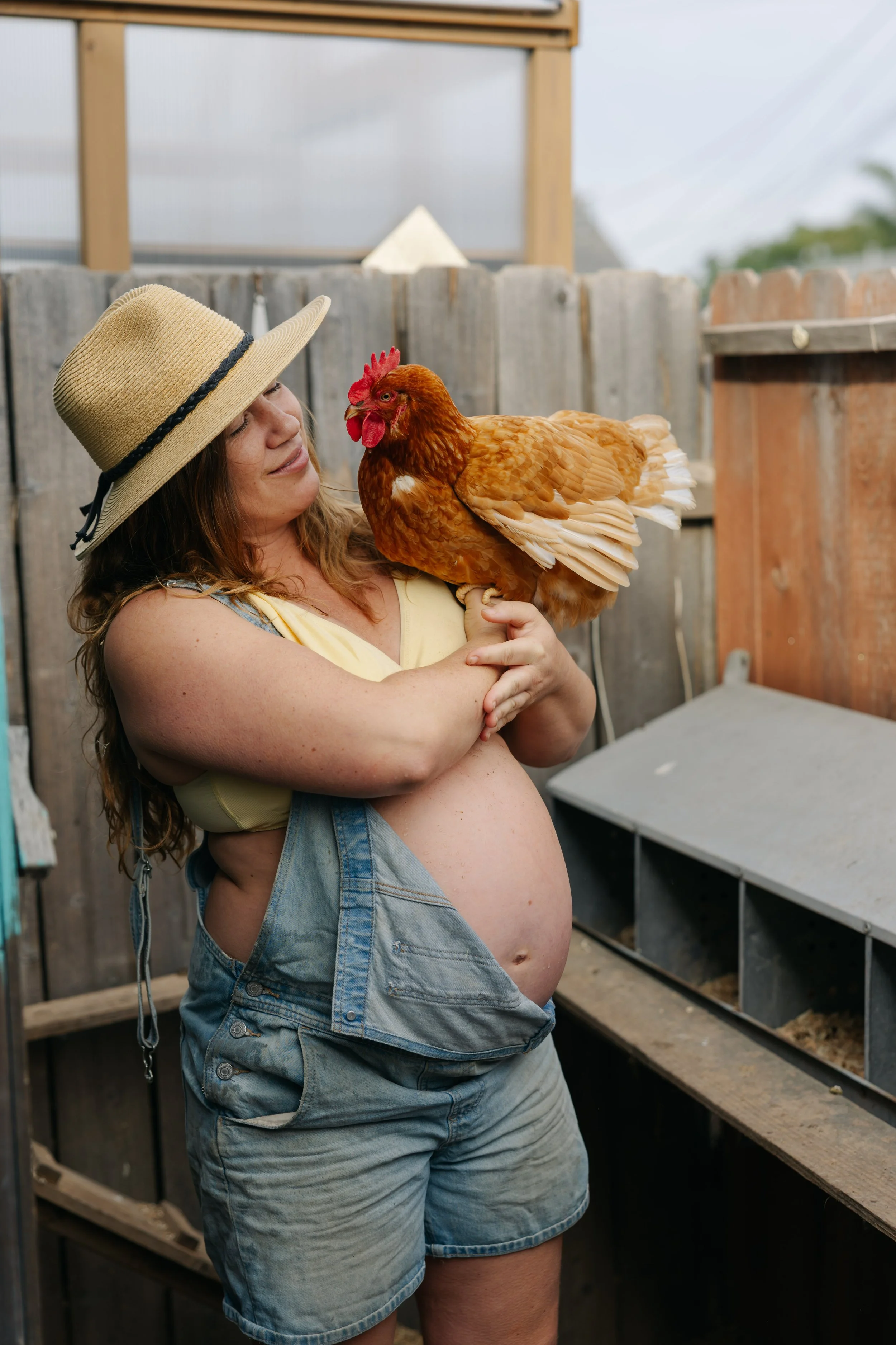 A pregnant woman in overalls holding a brown hen in a chicken coop.