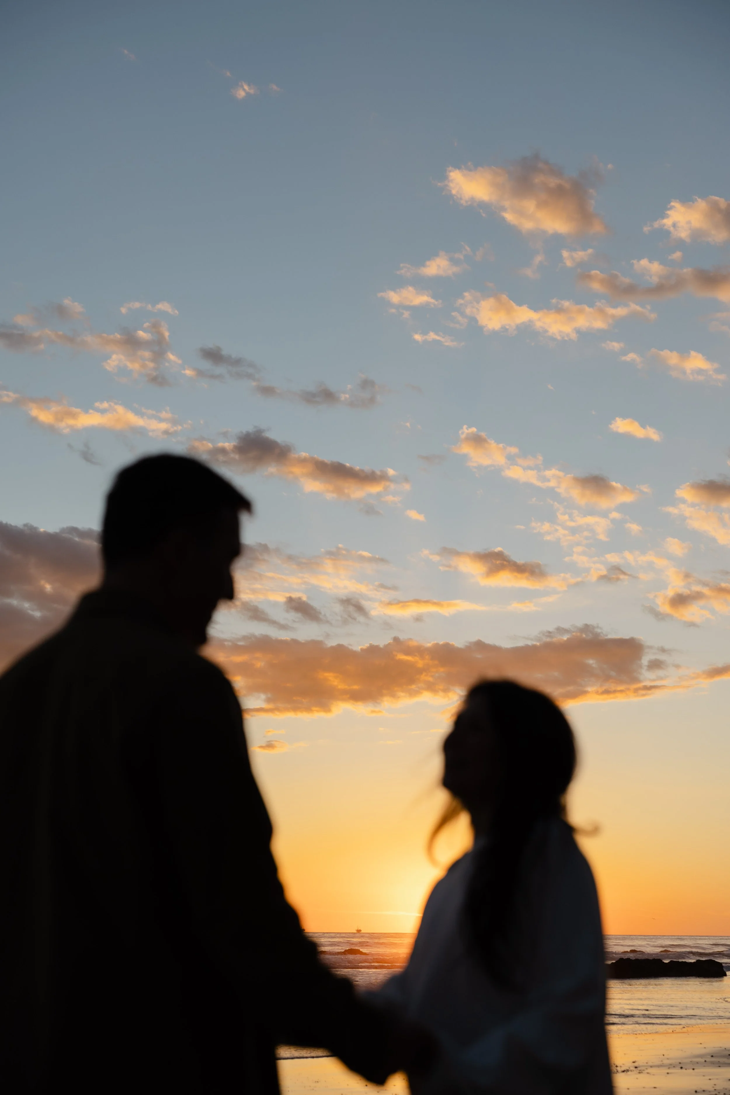 Silhouettes of a man and woman holding hands on a beach at sunset with clouds in the sky.