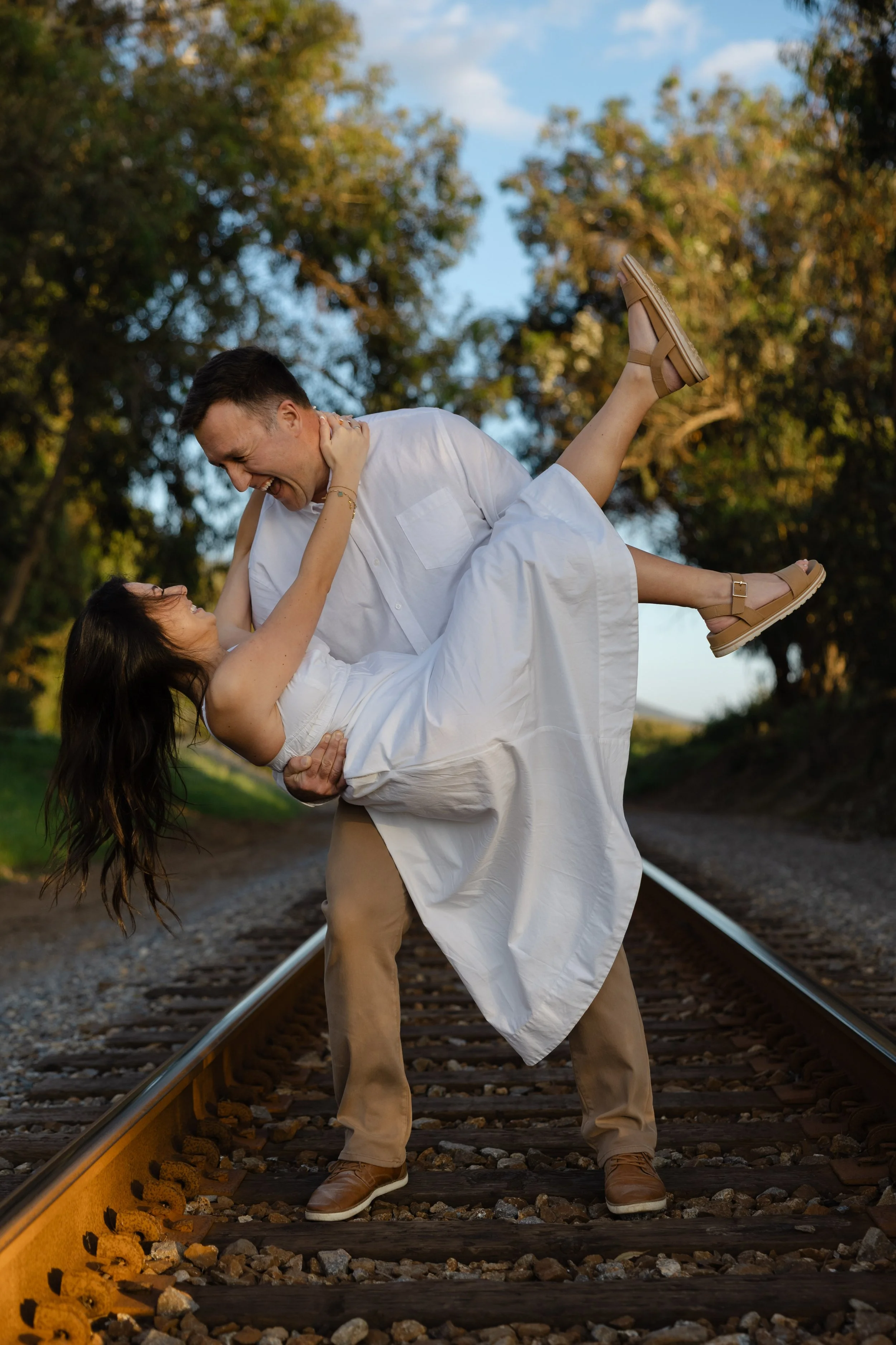 A man and woman are on train tracks, with the man holding the woman in a dip. They are smiling and laughing, surrounded by trees and a blue sky.