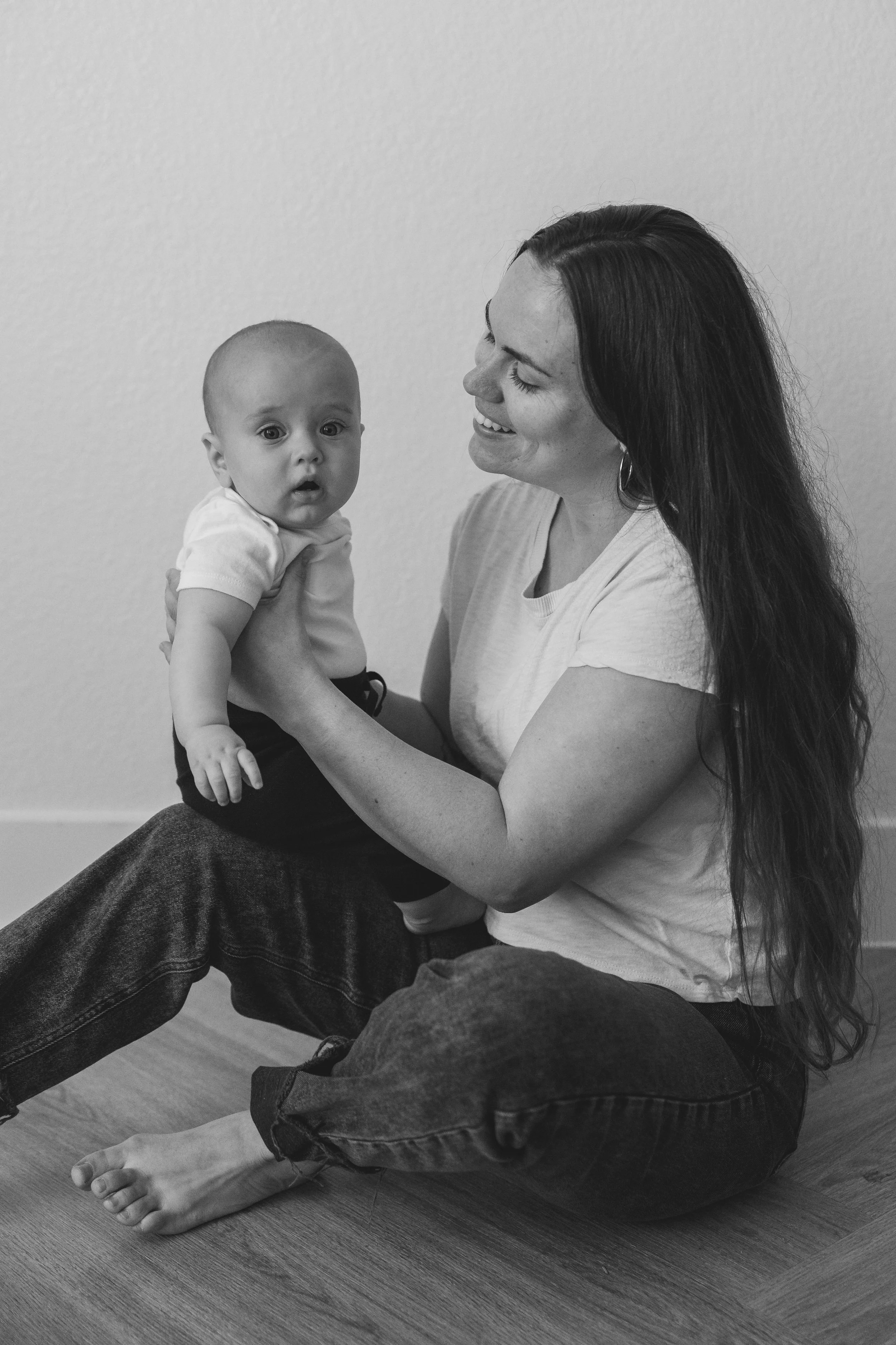 A woman with long dark hair sitting on the floor, holding a baby with a surprised expression, against a plain wall.