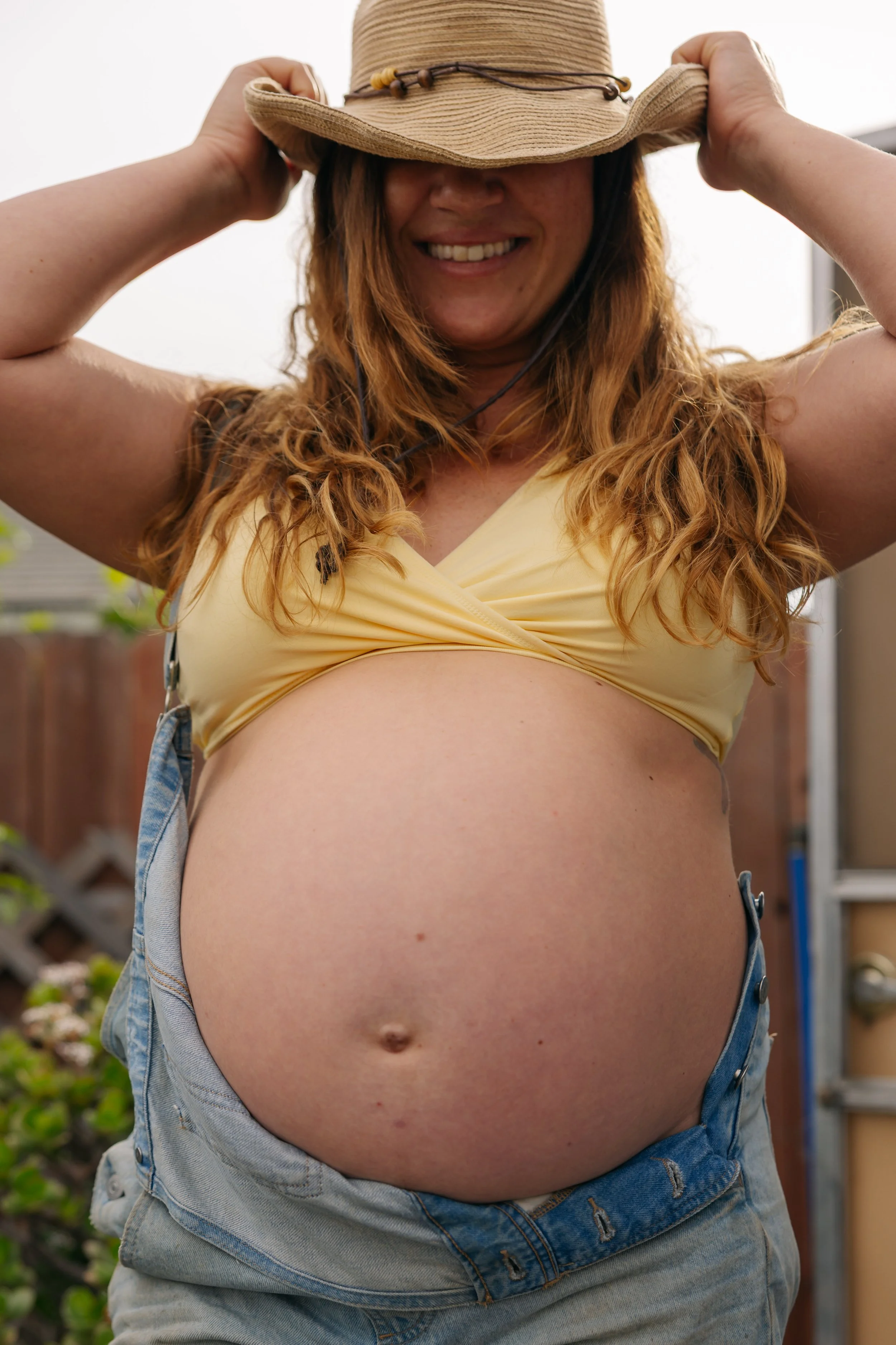 A pregnant woman wearing a yellow crop top, light blue jeans, and a brown hat, smiling while holding her hat.