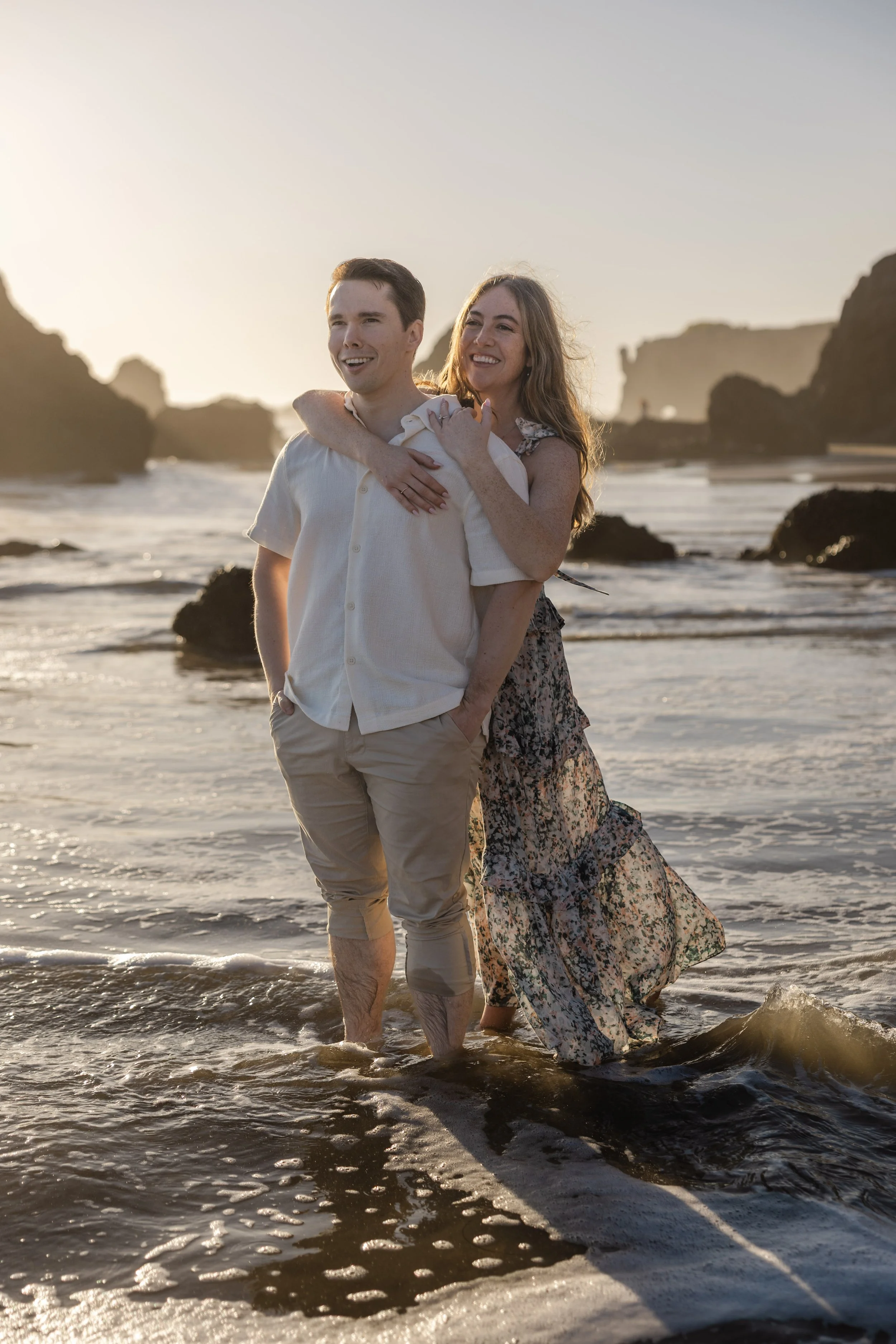 A smiling couple walking barefoot on a beach at sunset, with rocky formations in the background.