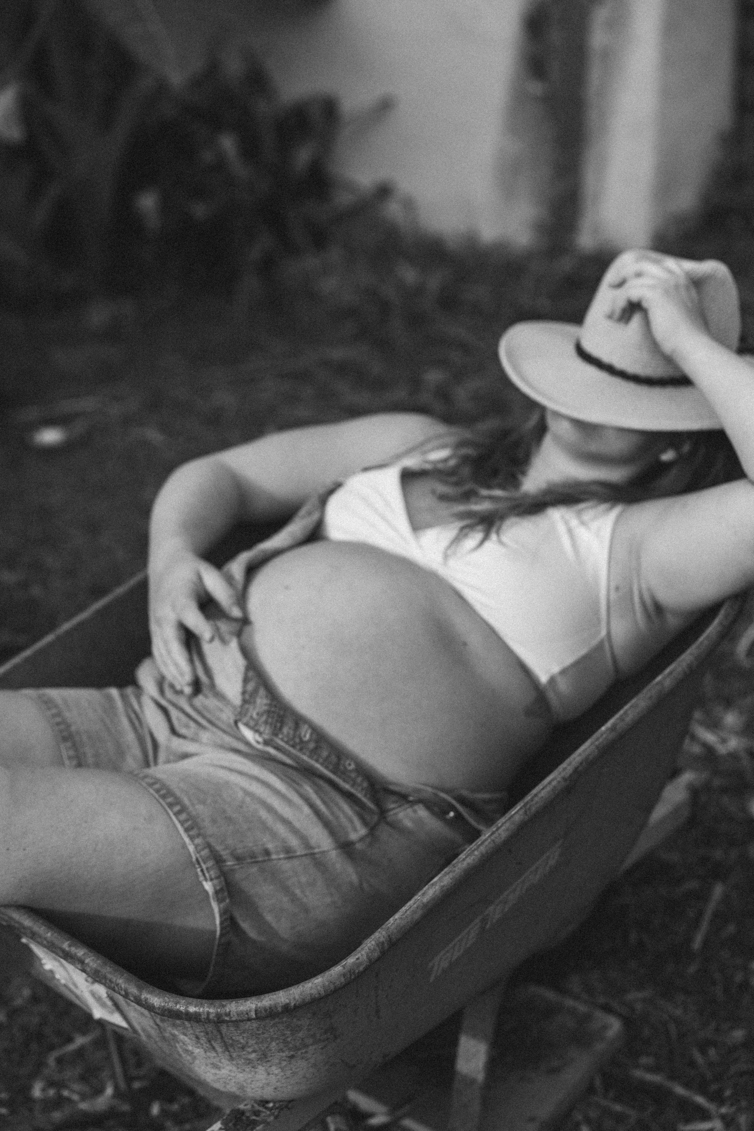 A woman lying in a wheelbarrow, wearing a hat and casual clothes, with her hand on her stomach.