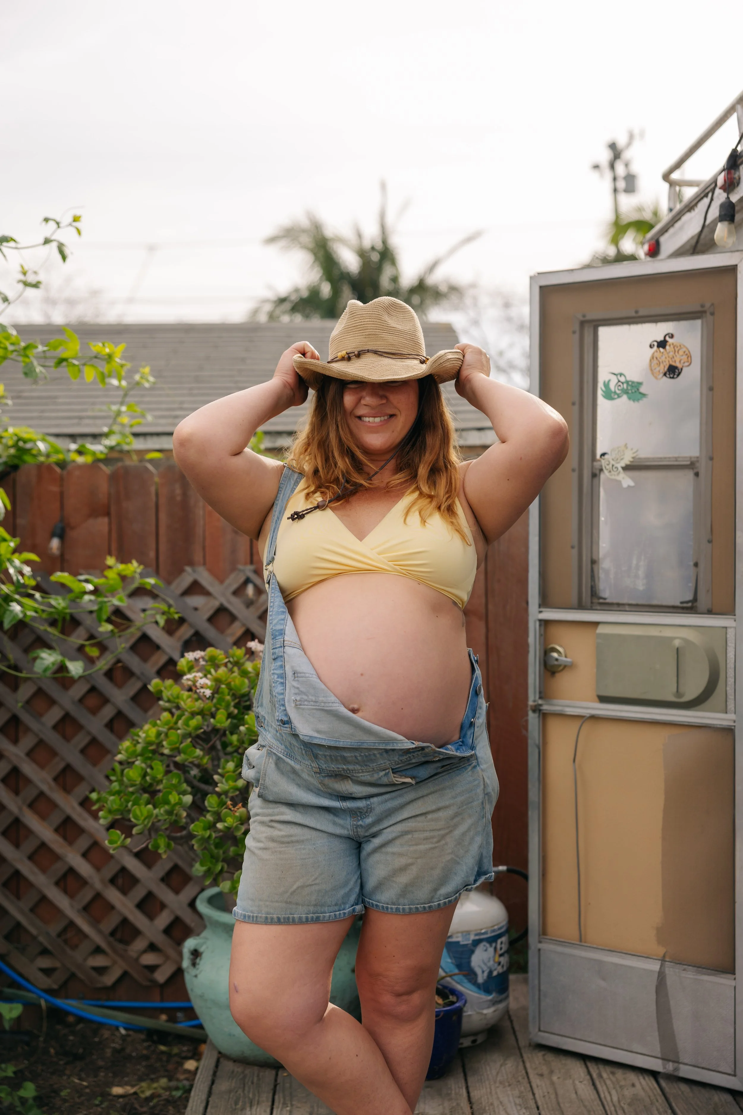 A pregnant woman standing outdoors, smiling, wearing a yellow crop top, denim shorts, and a wide-brimmed hat, with her hands adjusting her hat. She is standing in a backyard with a wooden fence, plants, and a small shed in the background.