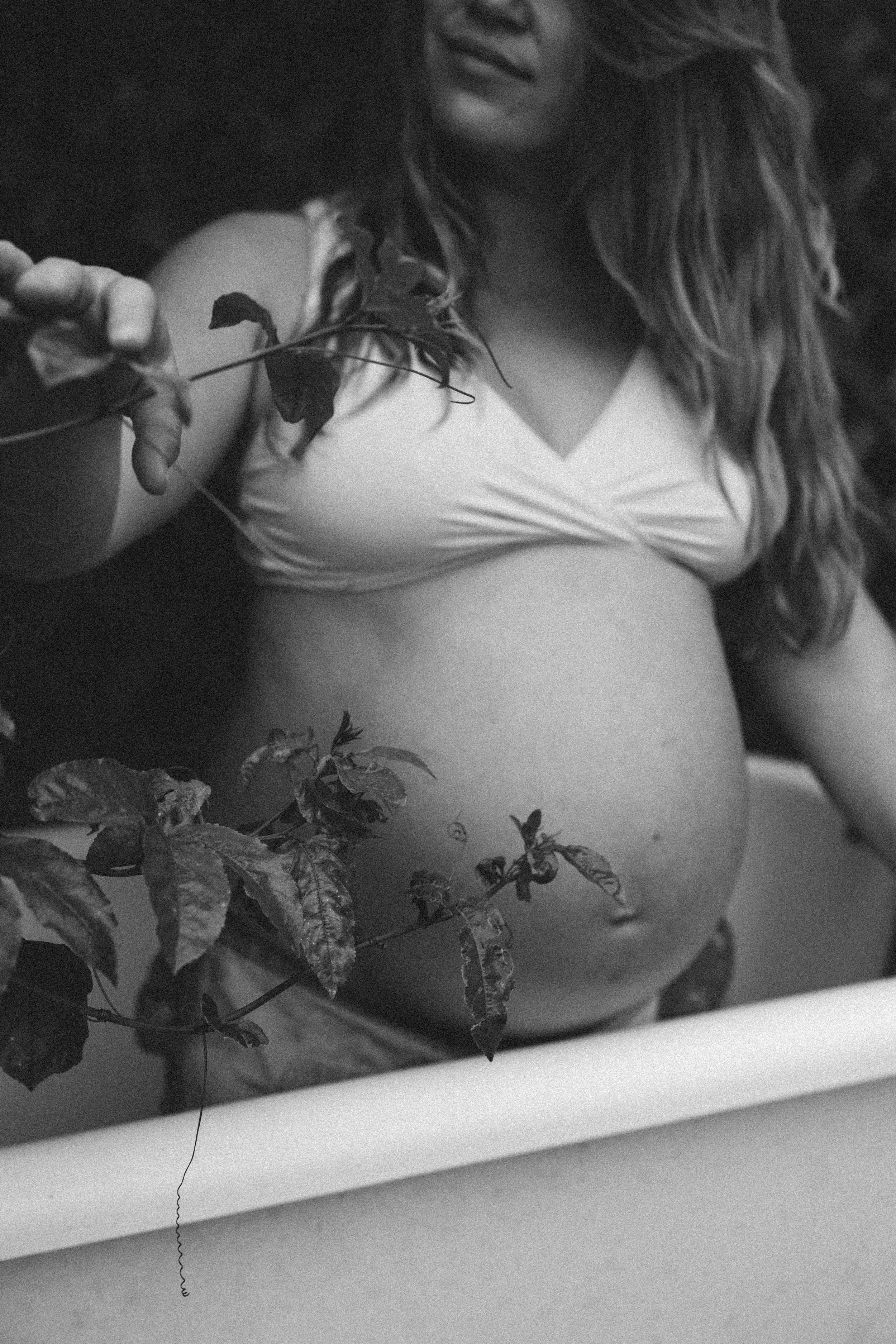 A pregnant woman with wavy hair holding a flower branch, wearing a white top, in black and white.