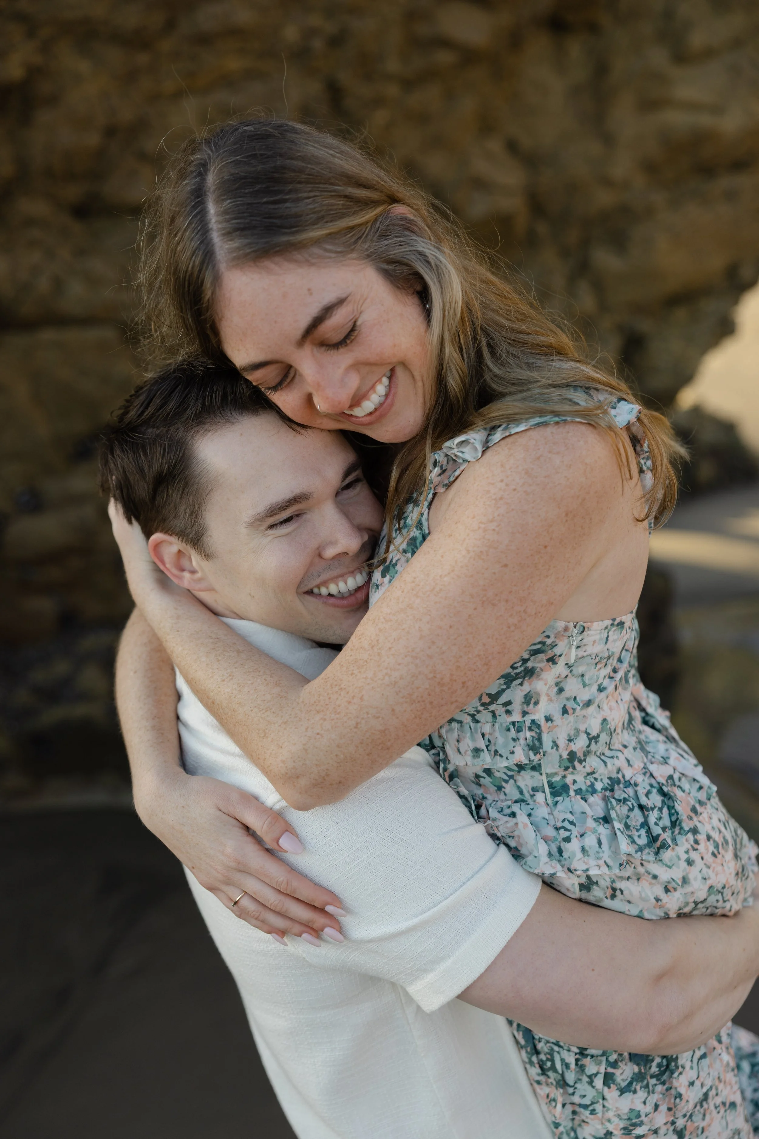 A woman with long hair and freckles hugging a smiling man with short dark hair in front of a natural rock background.