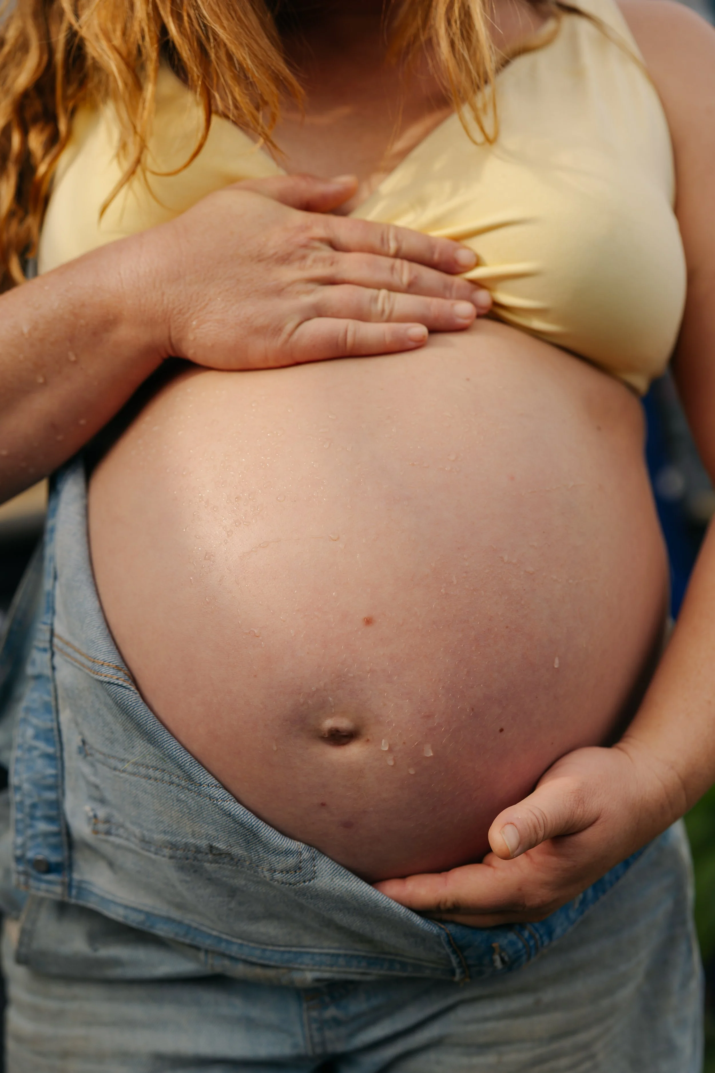 Close-up of a pregnant woman with her hand resting on her large belly, wearing a yellow top and denim shorts, with water droplets on her skin.