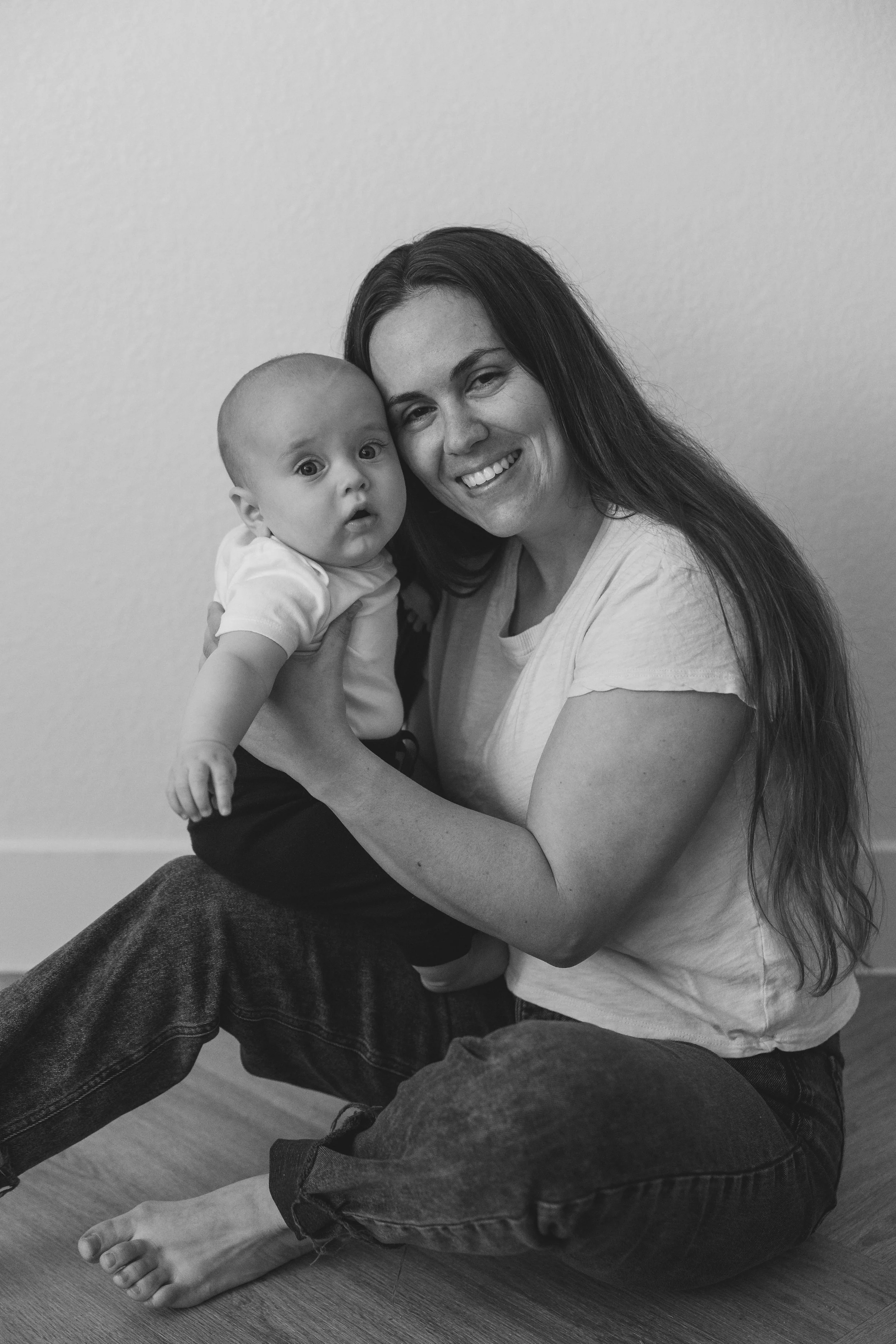A woman with long hair smiling while holding a baby with wide eyes and a curious expression, sitting on the floor against a plain wall.