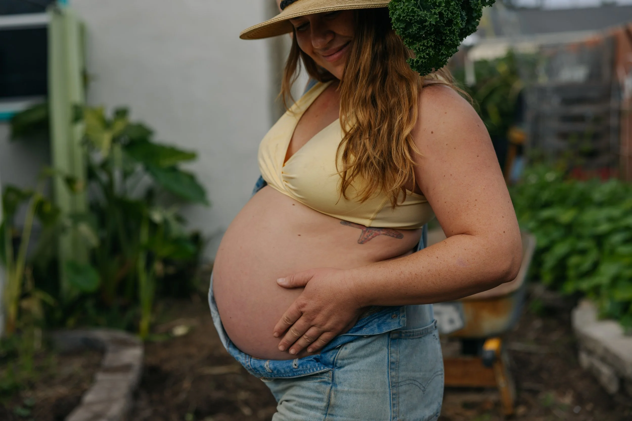 A pregnant woman with red hair, wearing a straw hat, yellow top, and denim shorts, standing outdoors in a garden, smiling and touching her belly.