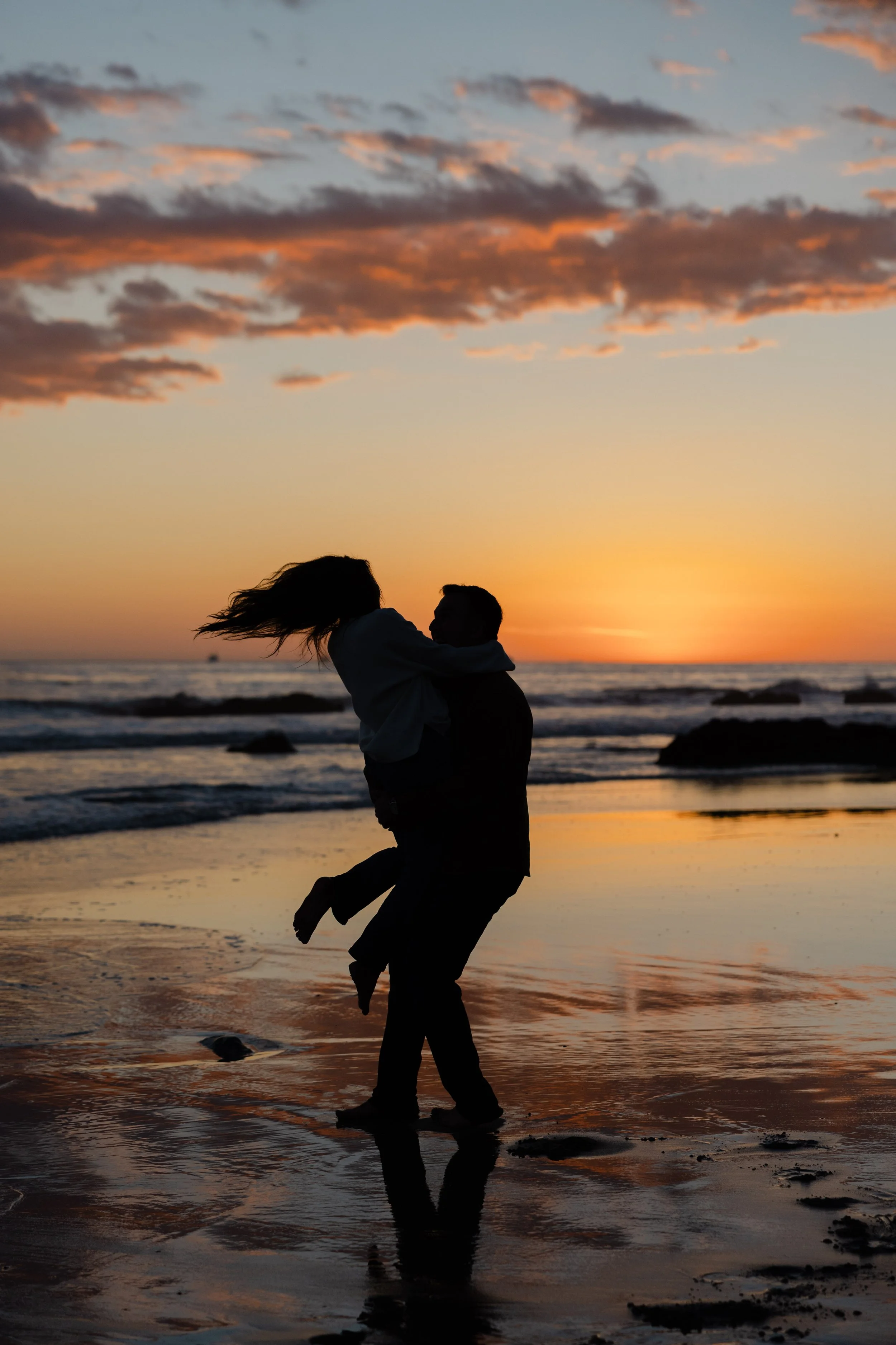 A silhouette of a man lifting a woman at the beach during sunset, with the ocean and colorful sky in the background.
