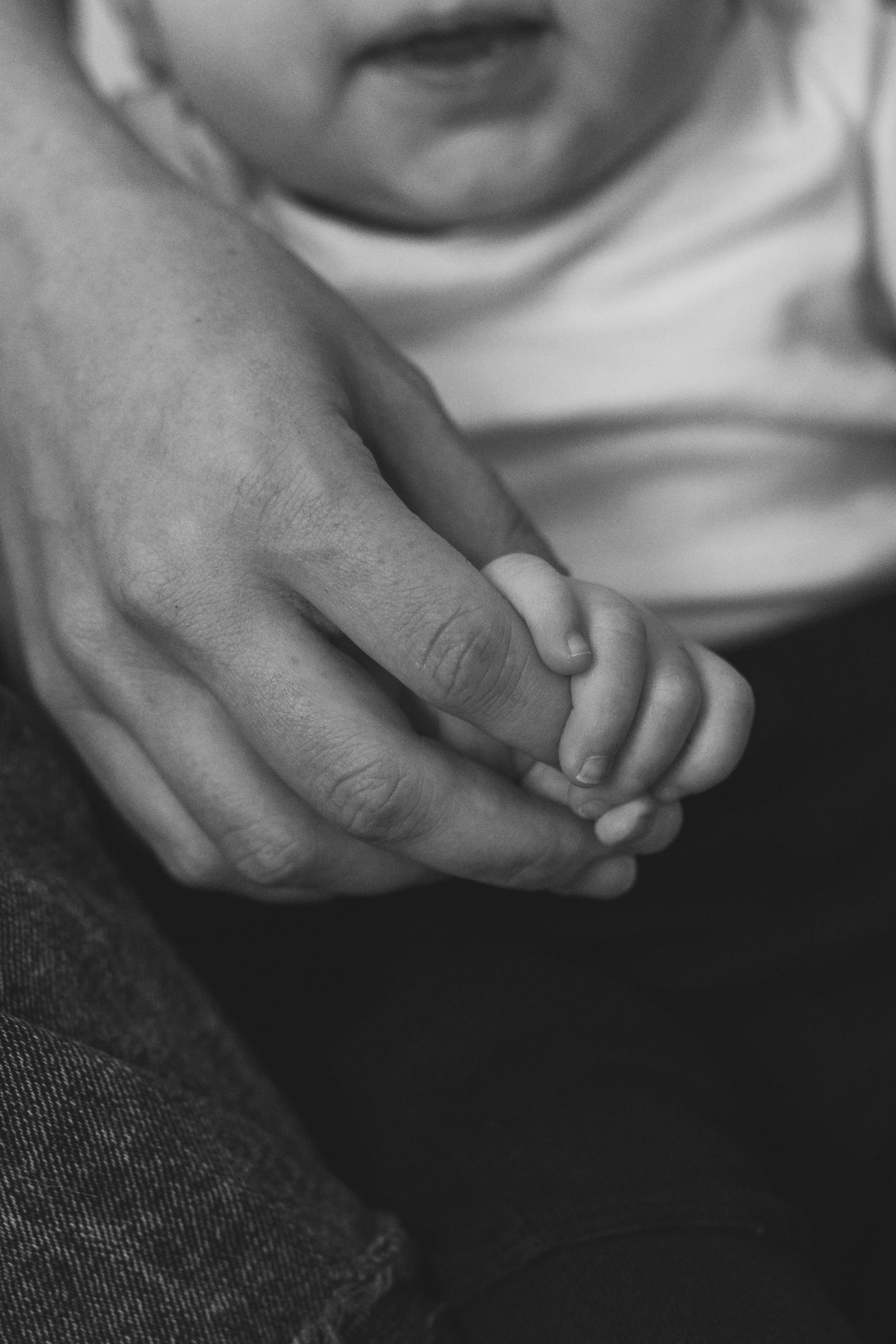 A close-up of an adult's hand holding a child's hand, both of them resting on their lap. The image is in black and white.