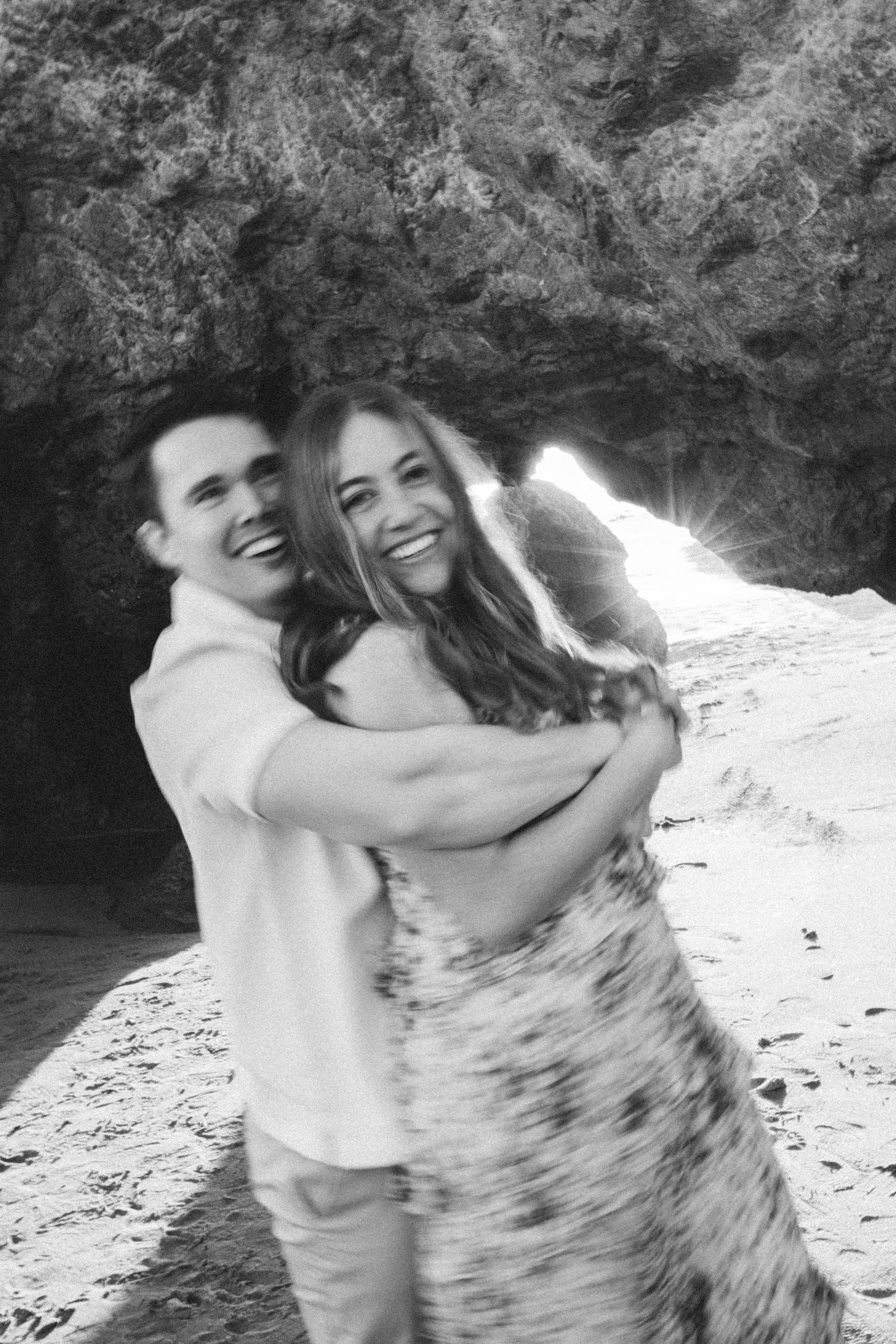 A happy couple hugging on a beach with a natural rock formation and an opening to the ocean in the background.