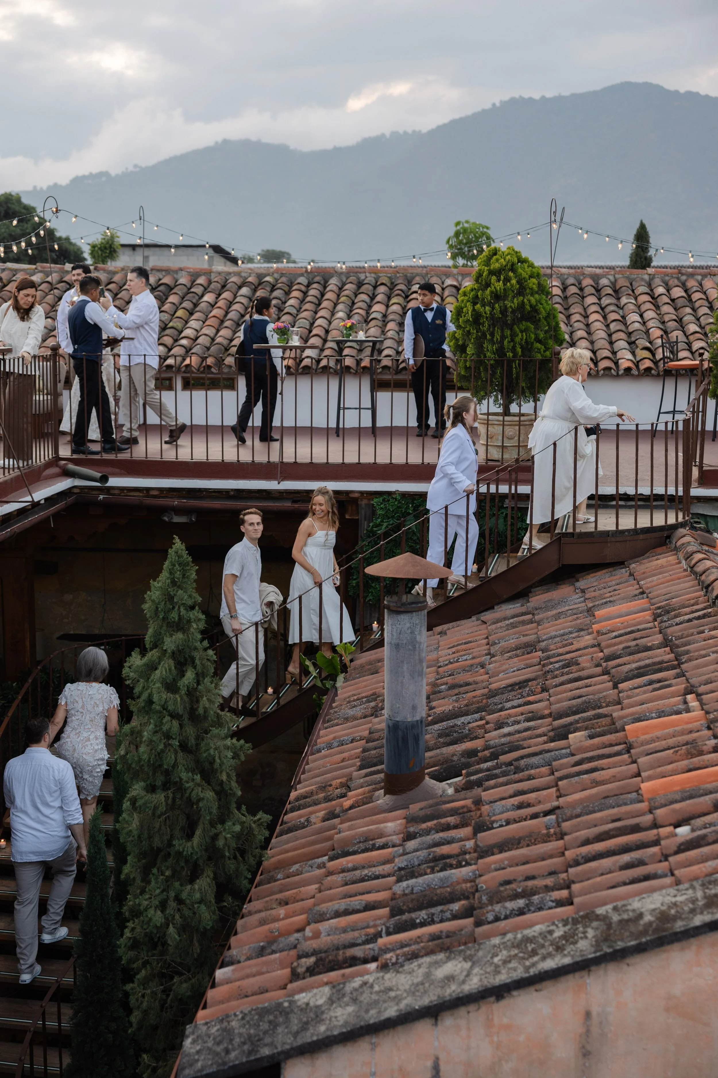 People dressed in white and dark clothing moving up and down an outdoor spiral staircase on a rooftop with a mountain landscape in the background.