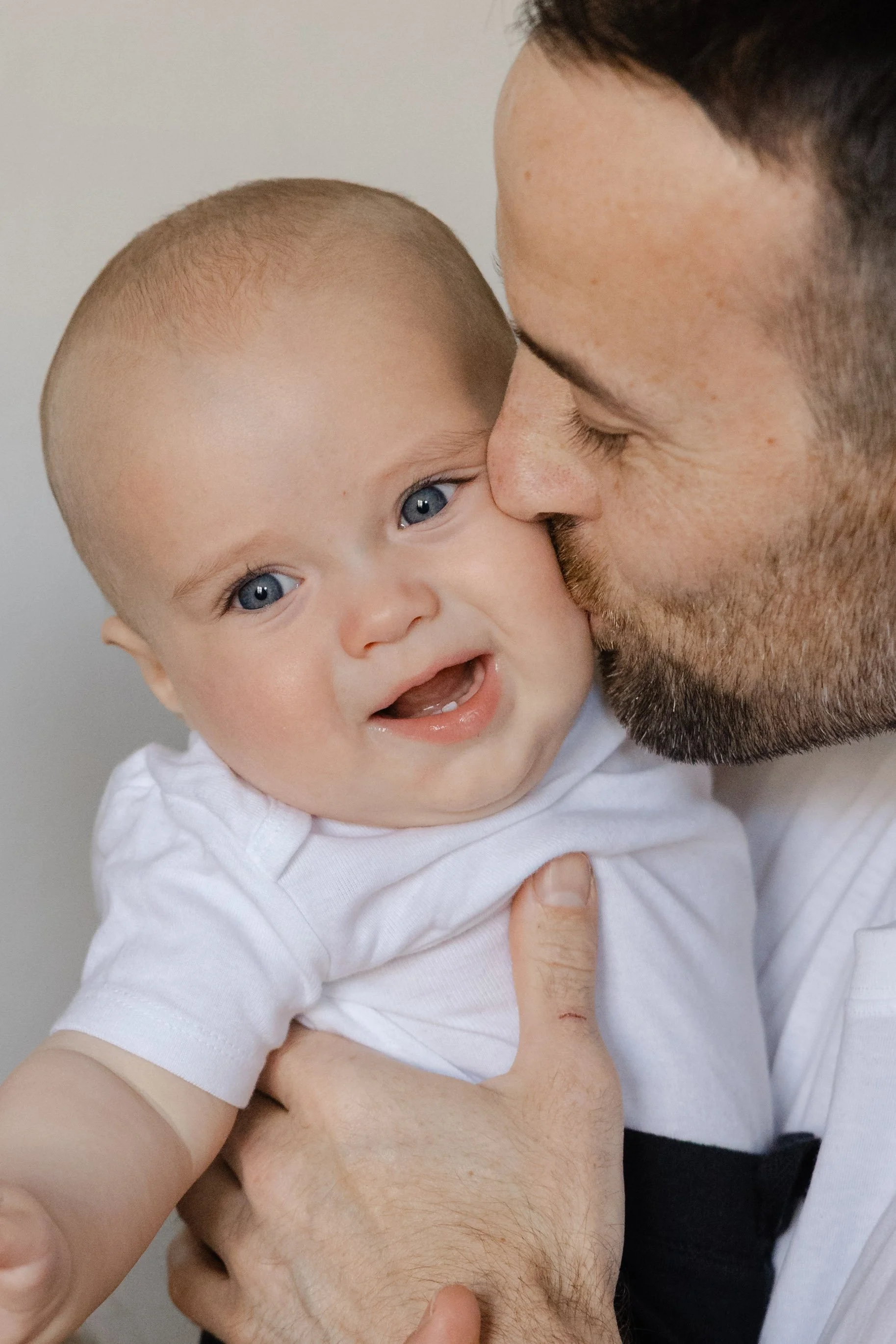 A man gives a kiss on the cheek to a crying baby with blue eyes, wearing a white shirt, against a plain background.