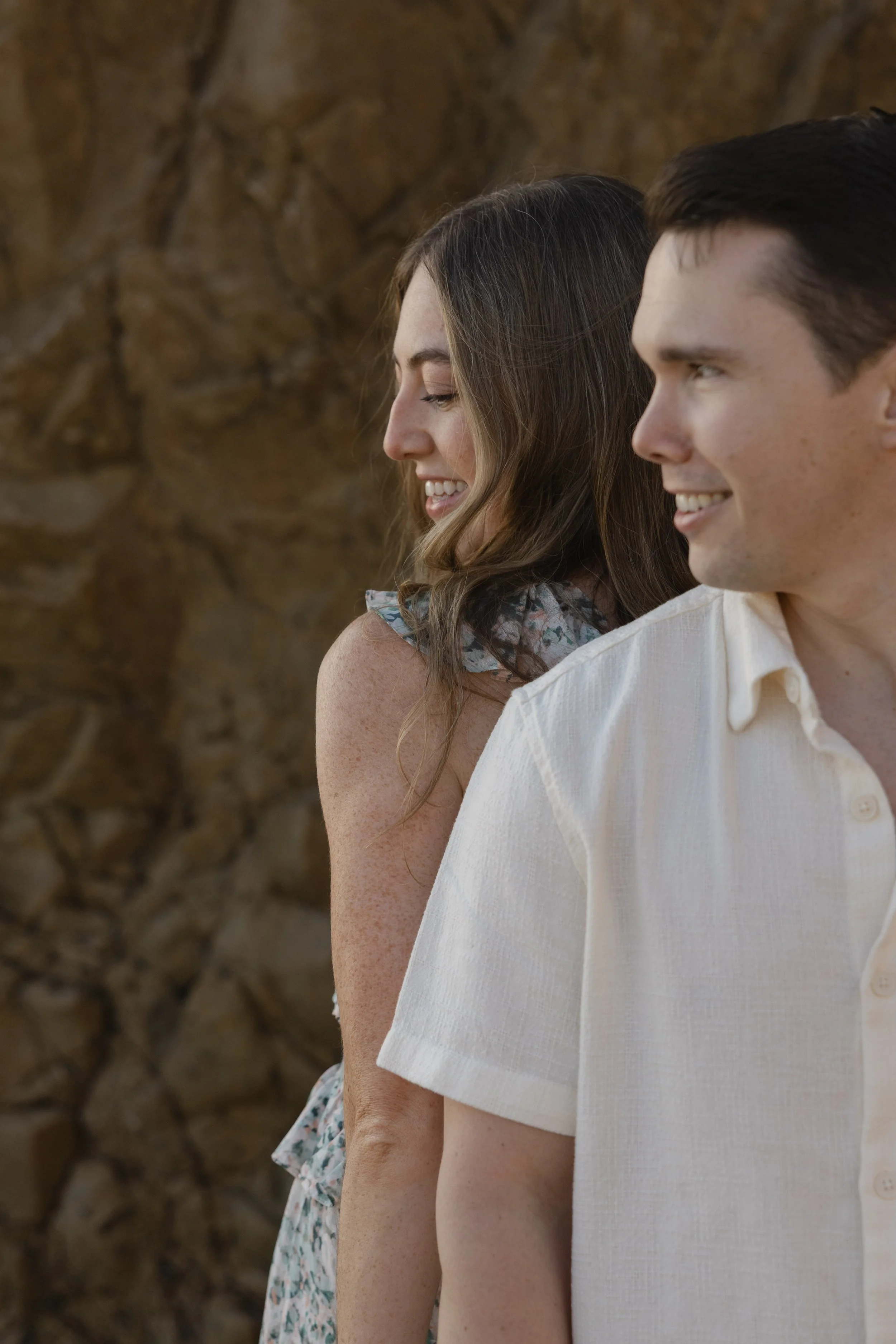 A young woman with long, wavy brown hair and a young man with short dark hair standing outdoors, back to back, smiling gently with a rocky background.
