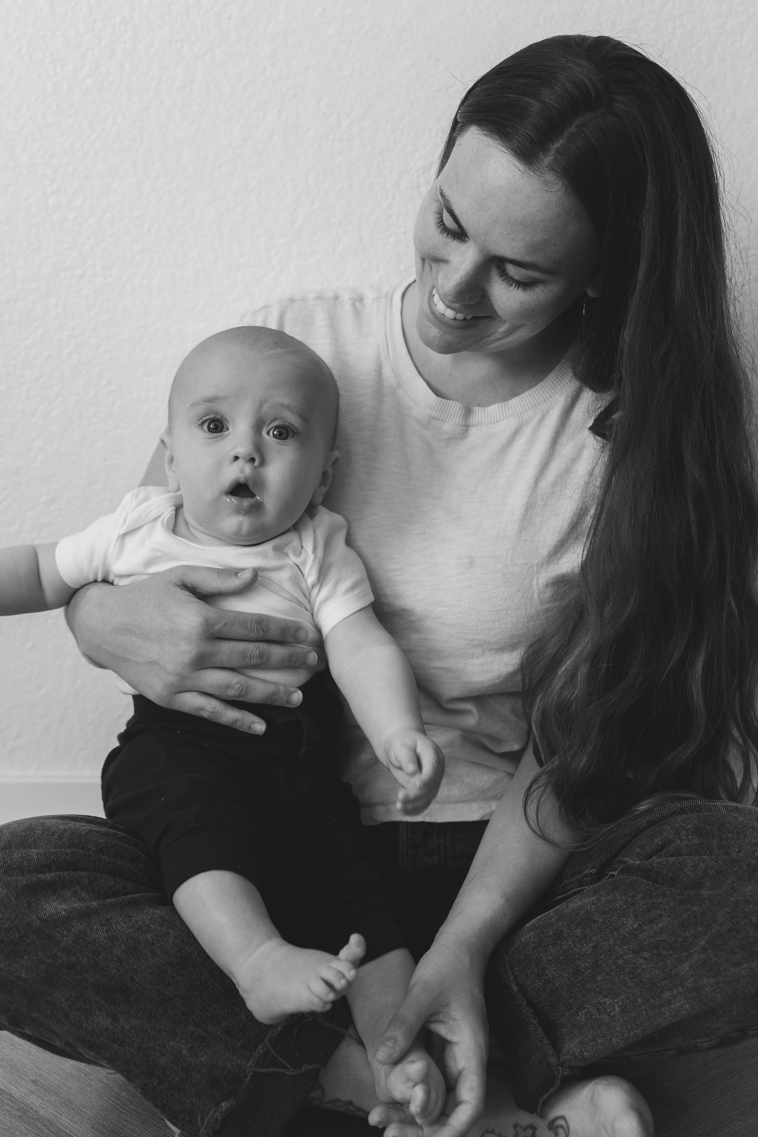 A woman holding a surprised baby on her lap, smiling and looking down.