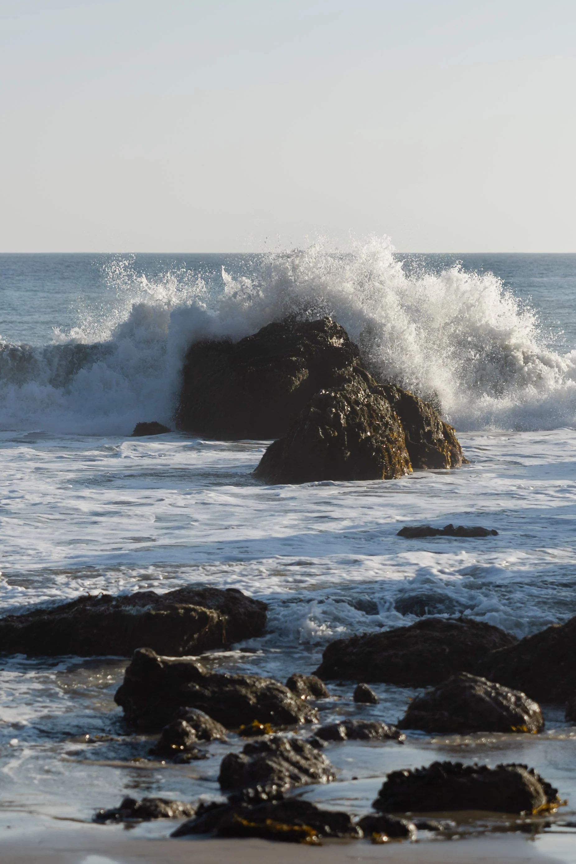 Waves crashing against large rocks on a beach with a clear sky in the background.