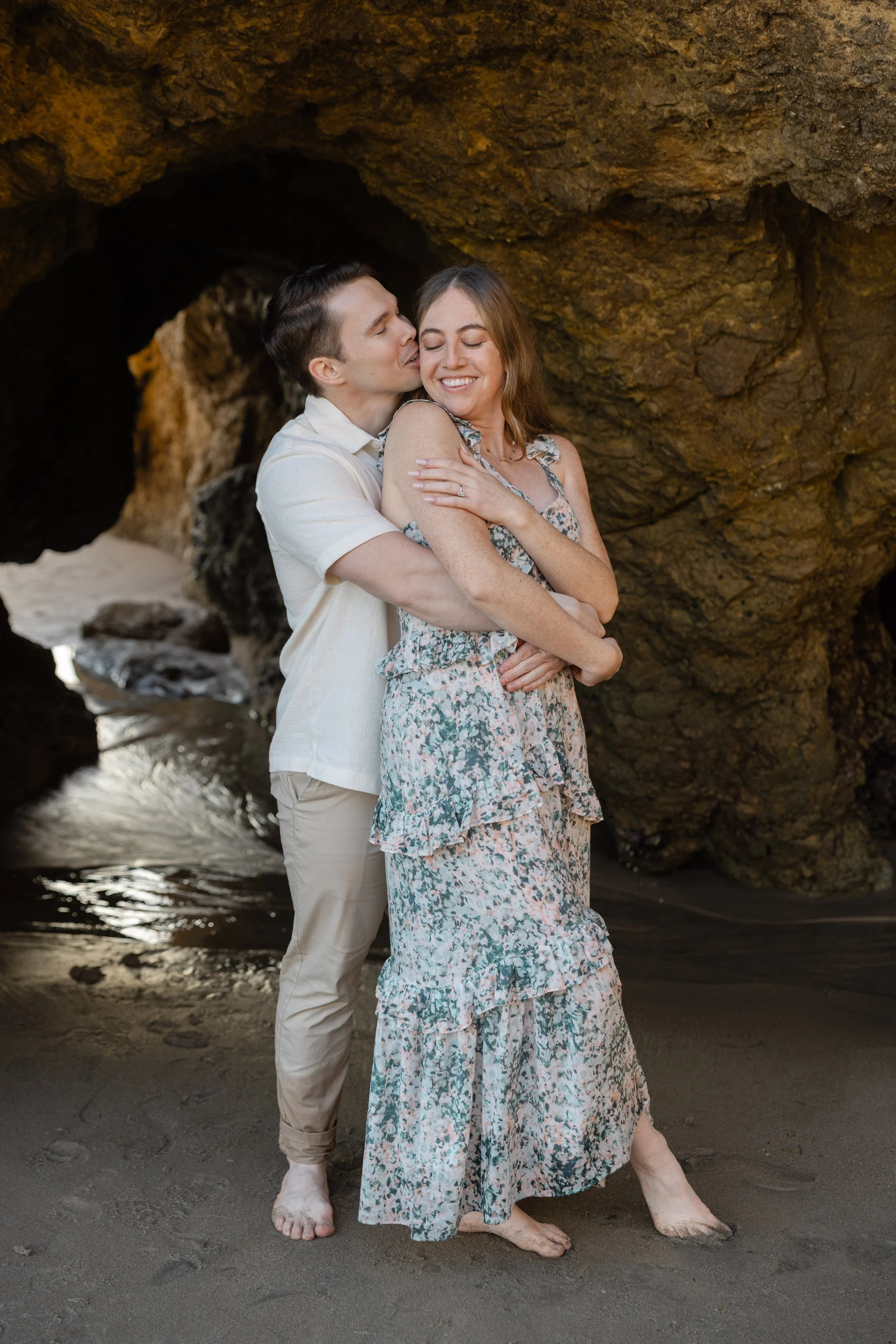 A couple standing on a sandy beach under a large rock formation, embracing each other and smiling, with the man kissing the woman on the cheek.