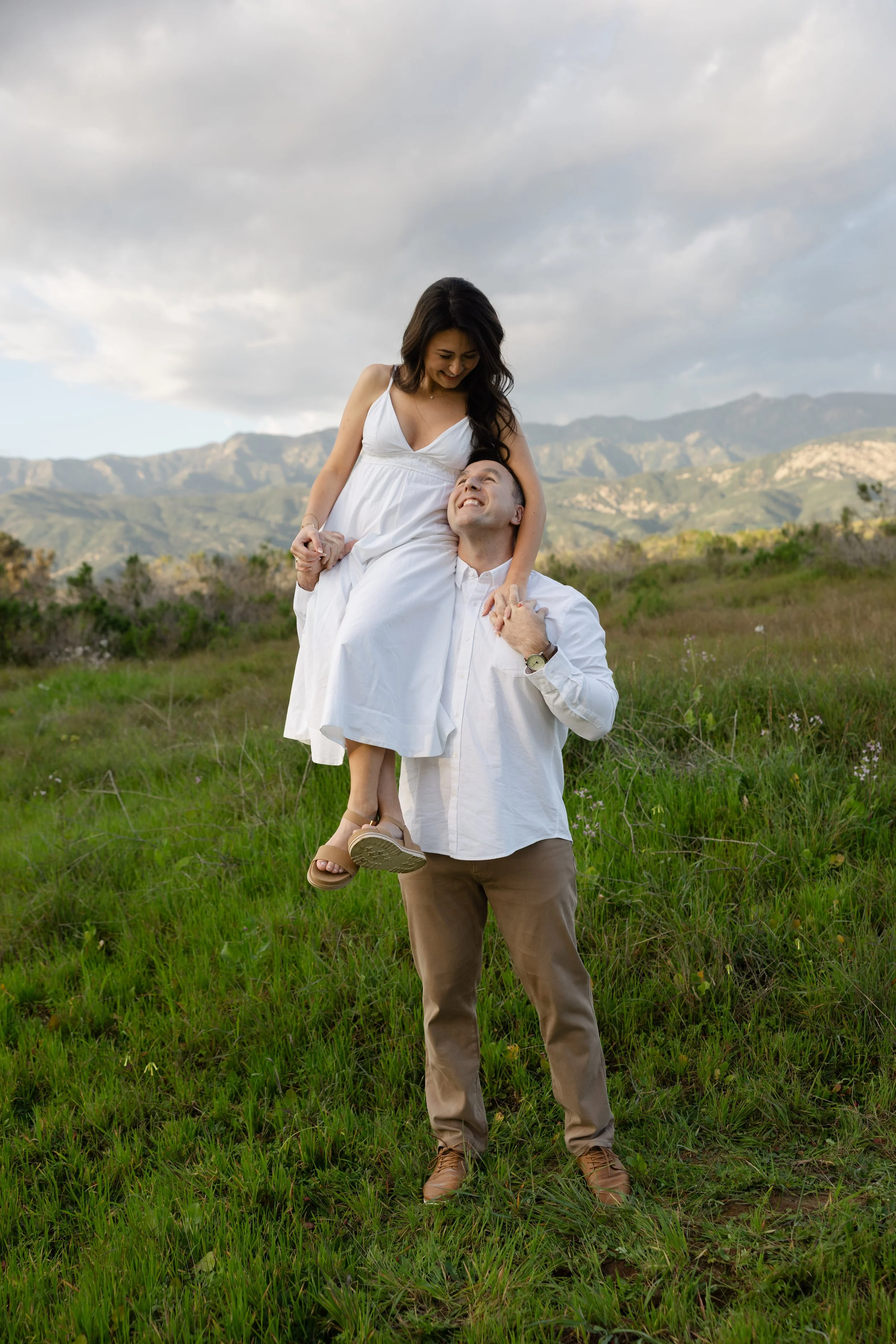 A couple outdoors, the man lifting the woman, both smiling, with mountains and cloudy sky in the background.