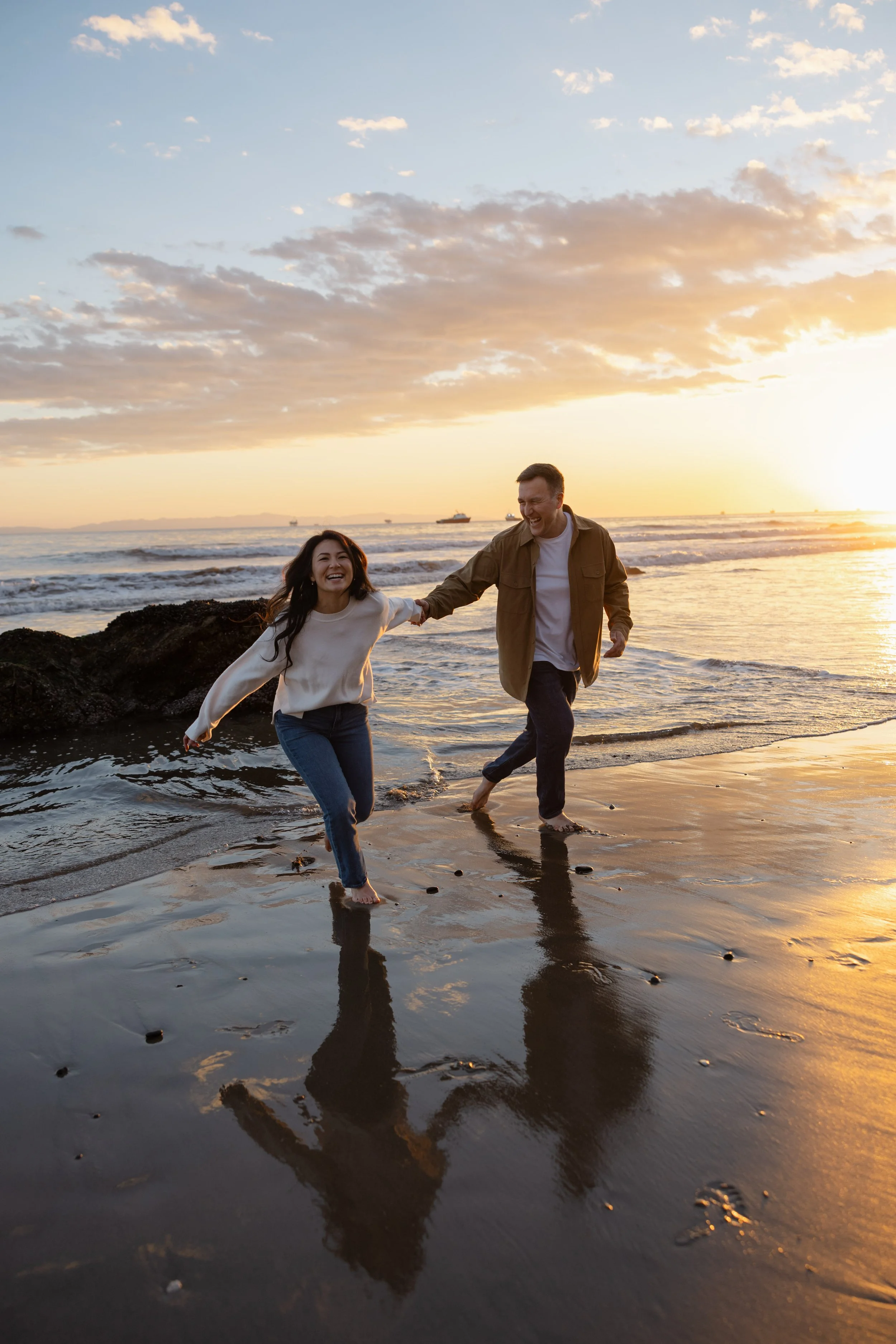 A couple running and playing in shallow ocean water at sunset on the beach, holding hands and smiling.