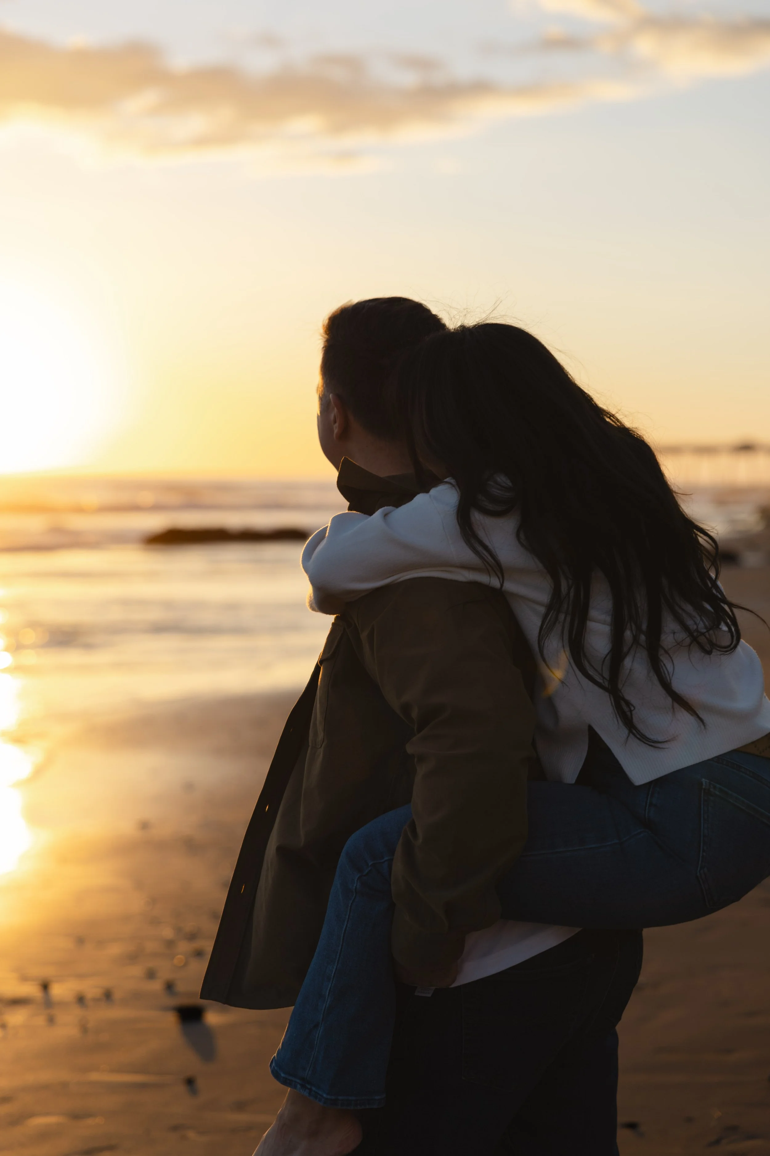 A couple at sunset on the beach, one person carrying the other on their back, with the ocean and sky in the background.