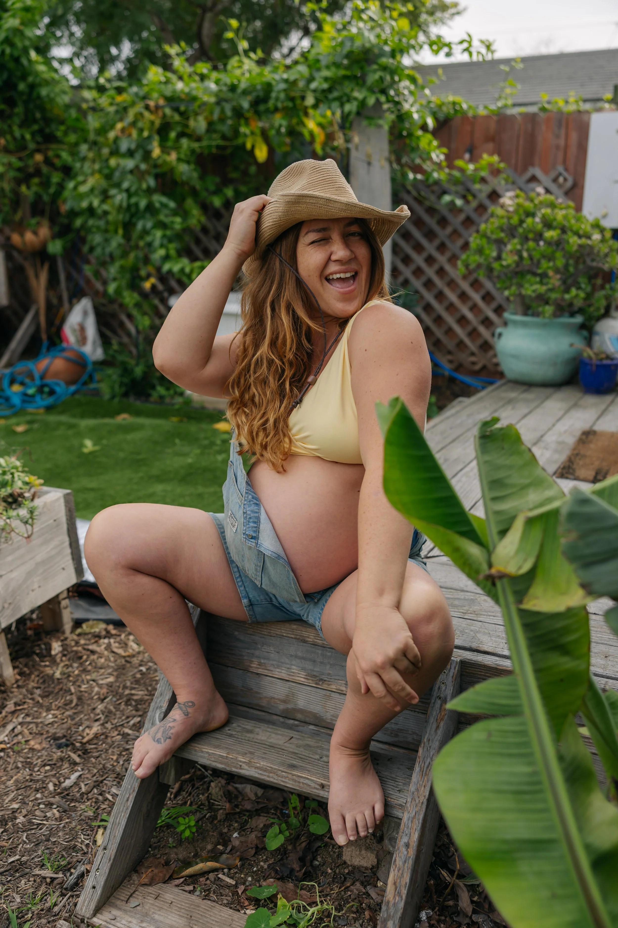 A pregnant woman with long curly hair, wearing a tan hat, yellow top, and denim shorts, sitting on a small wooden step outdoors, smiling and holding her hat, surrounded by greenery and garden decor.