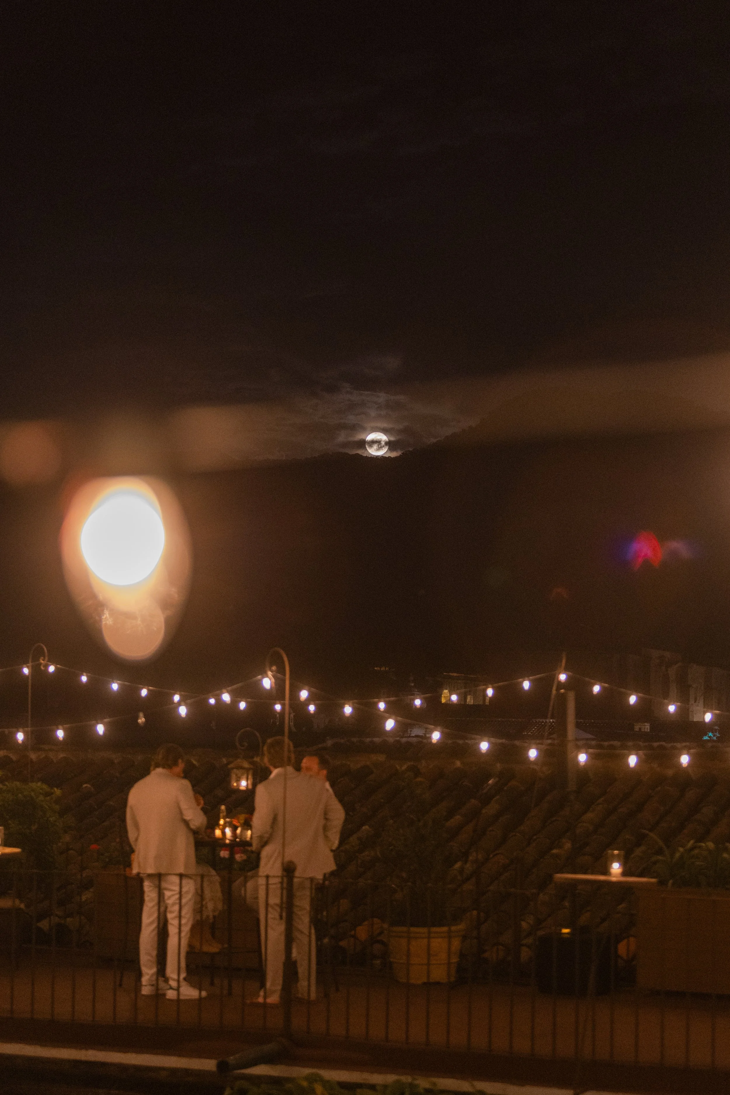 Night scene showing three people in white suits gathered outside, with string lights overhead, a mountain with the moon partially covered by clouds in the background, and lens flare artifacts.