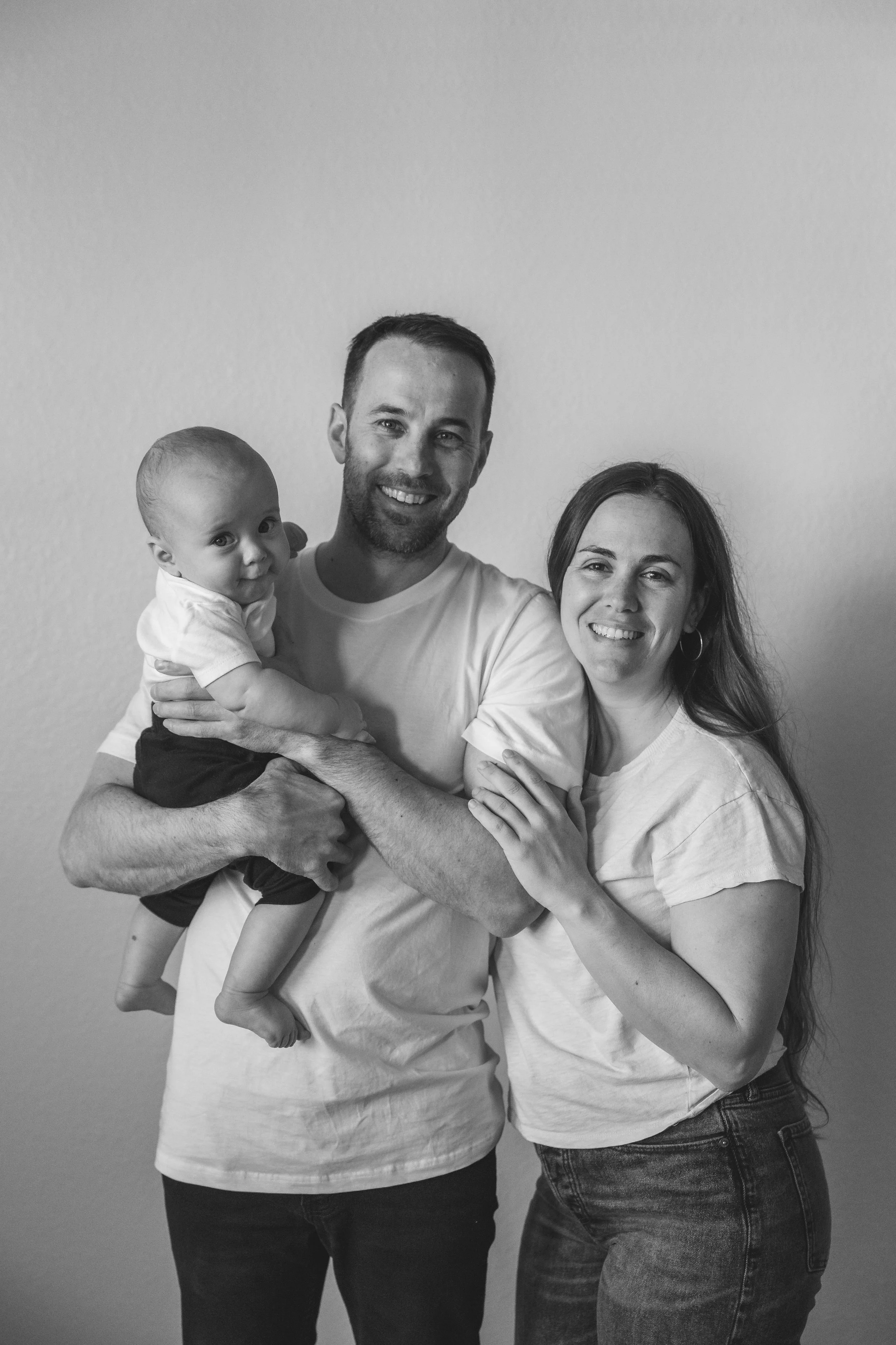 A smiling family of three: a man holding a baby boy in his arms, and a woman standing next to them, all in casual white T-shirts against a plain wall.