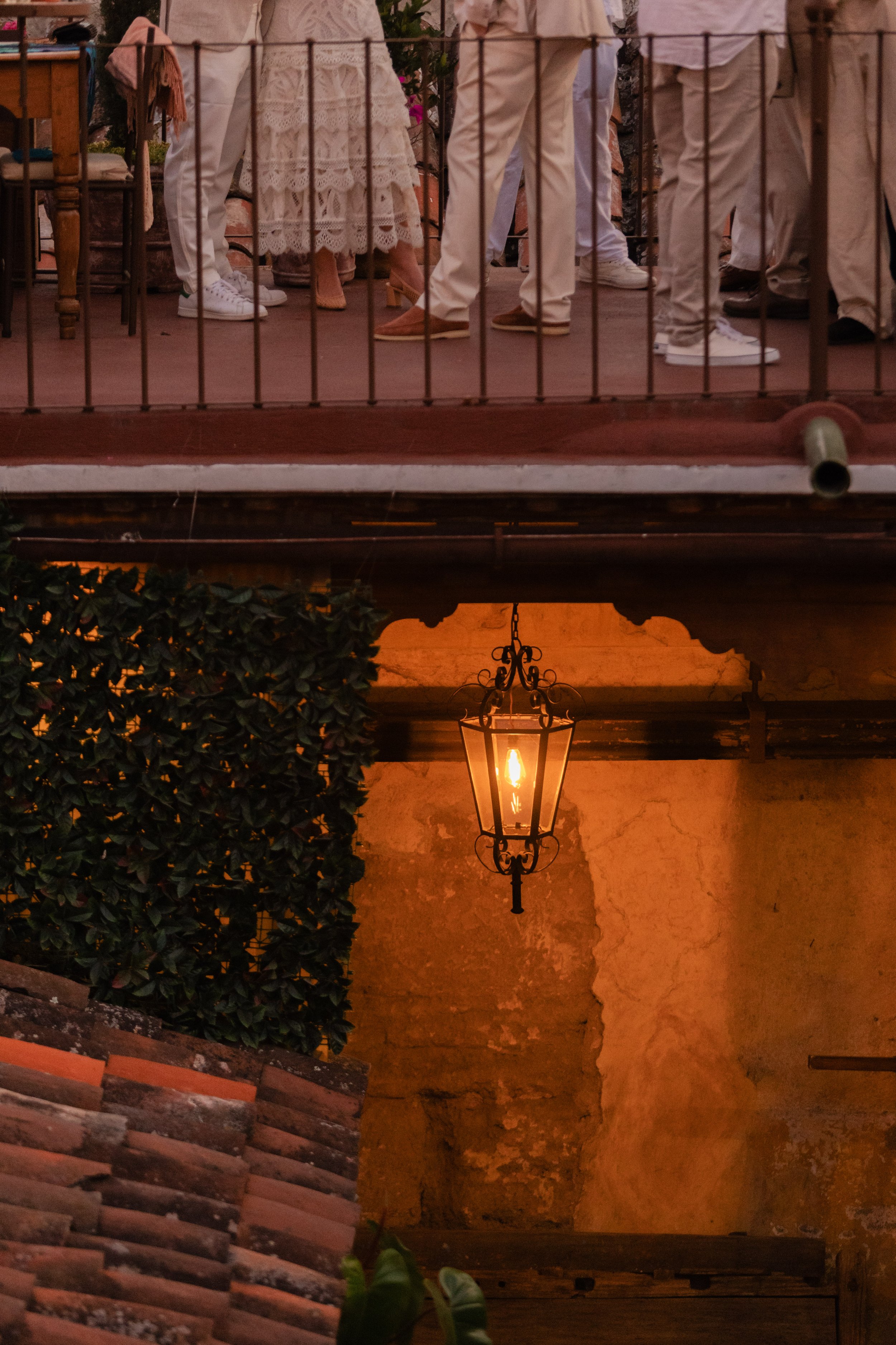 People dressed in white at a wedding ceremony on a balcony with a wrought iron railing, with a warm-colored wall and hanging lantern below.