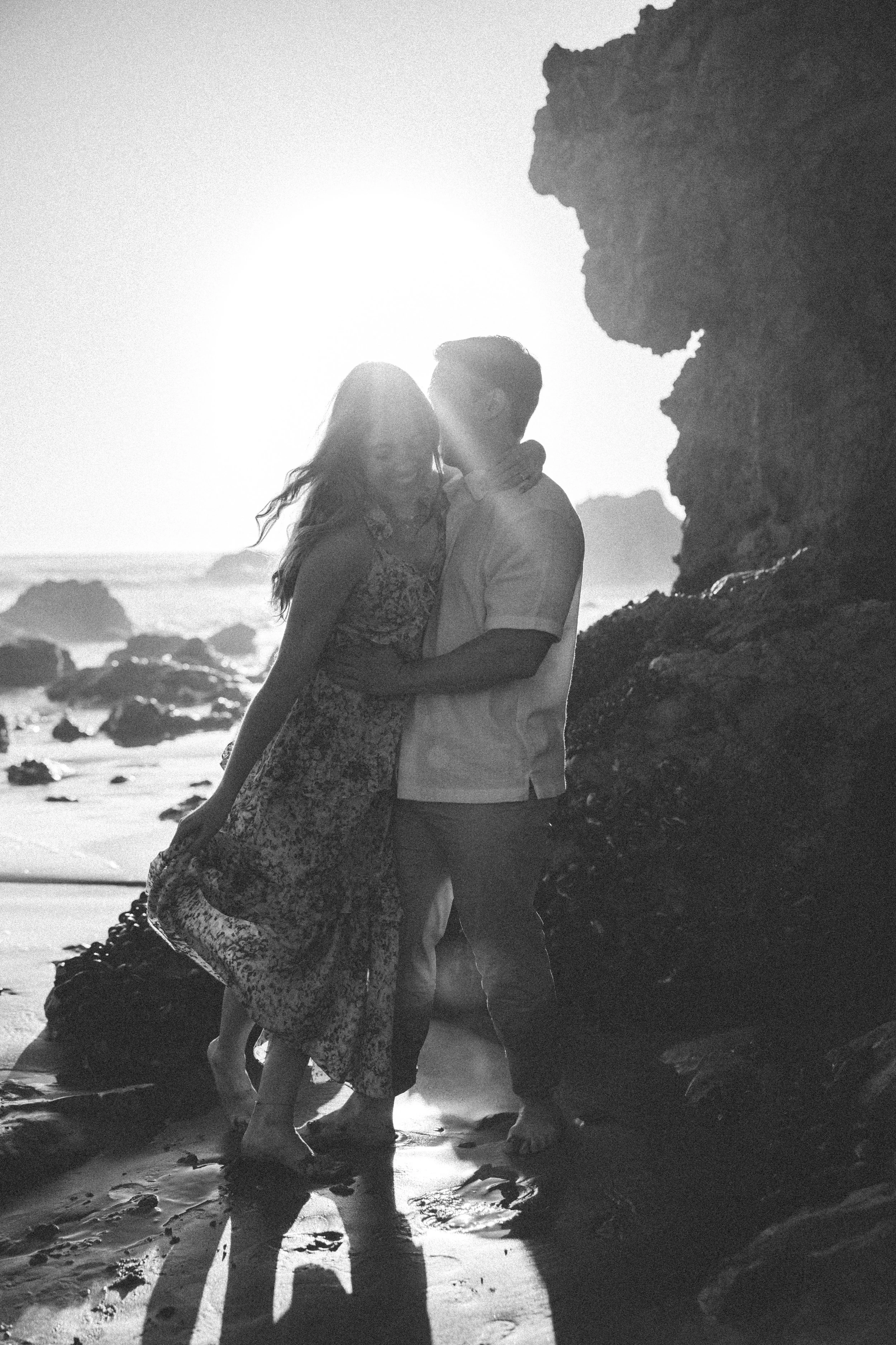 A black and white photo of a couple embracing on a beach near rocks during sunset.